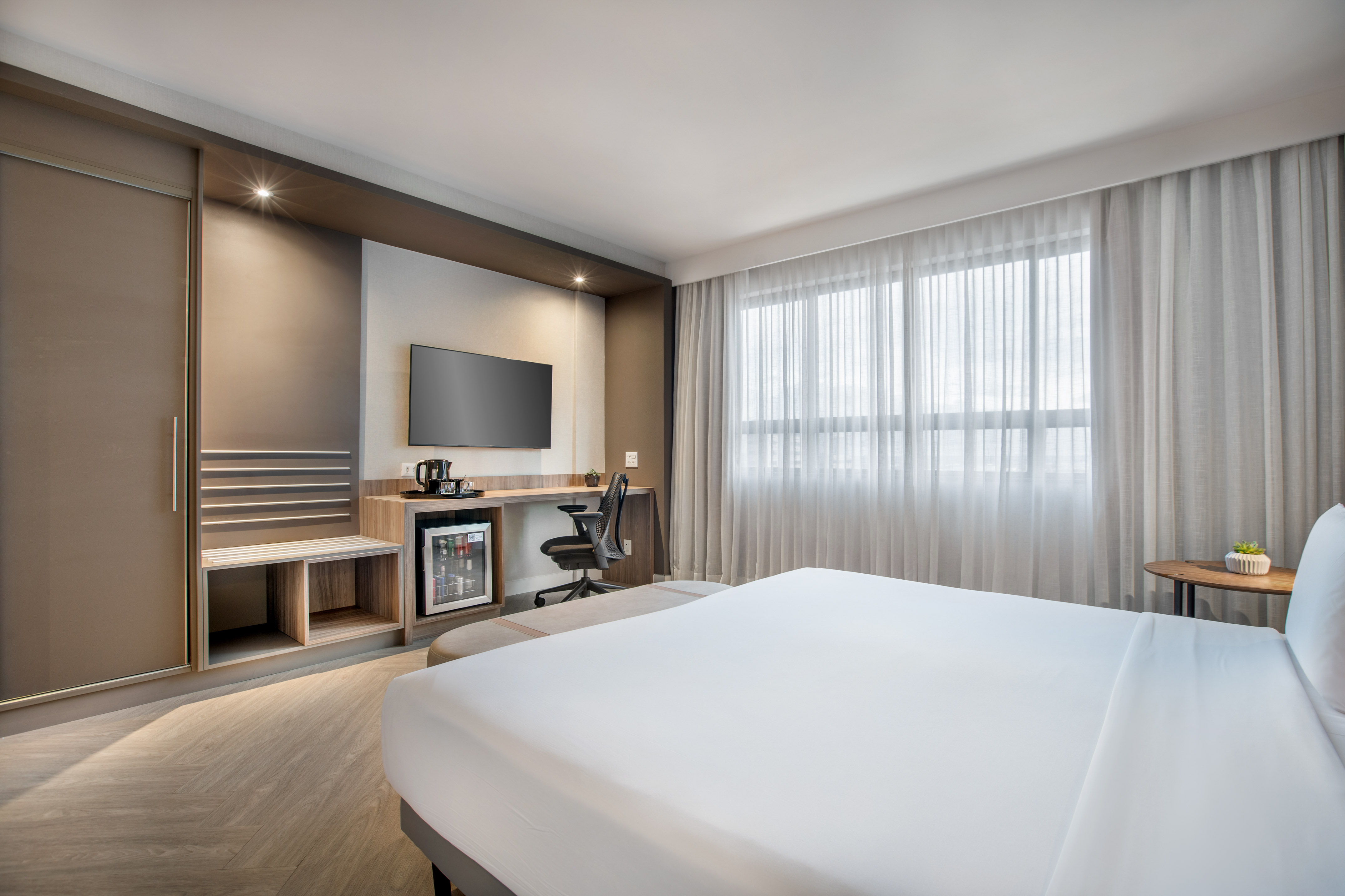 Modern hotel room with bed, desk, TV, minibar, and large window.