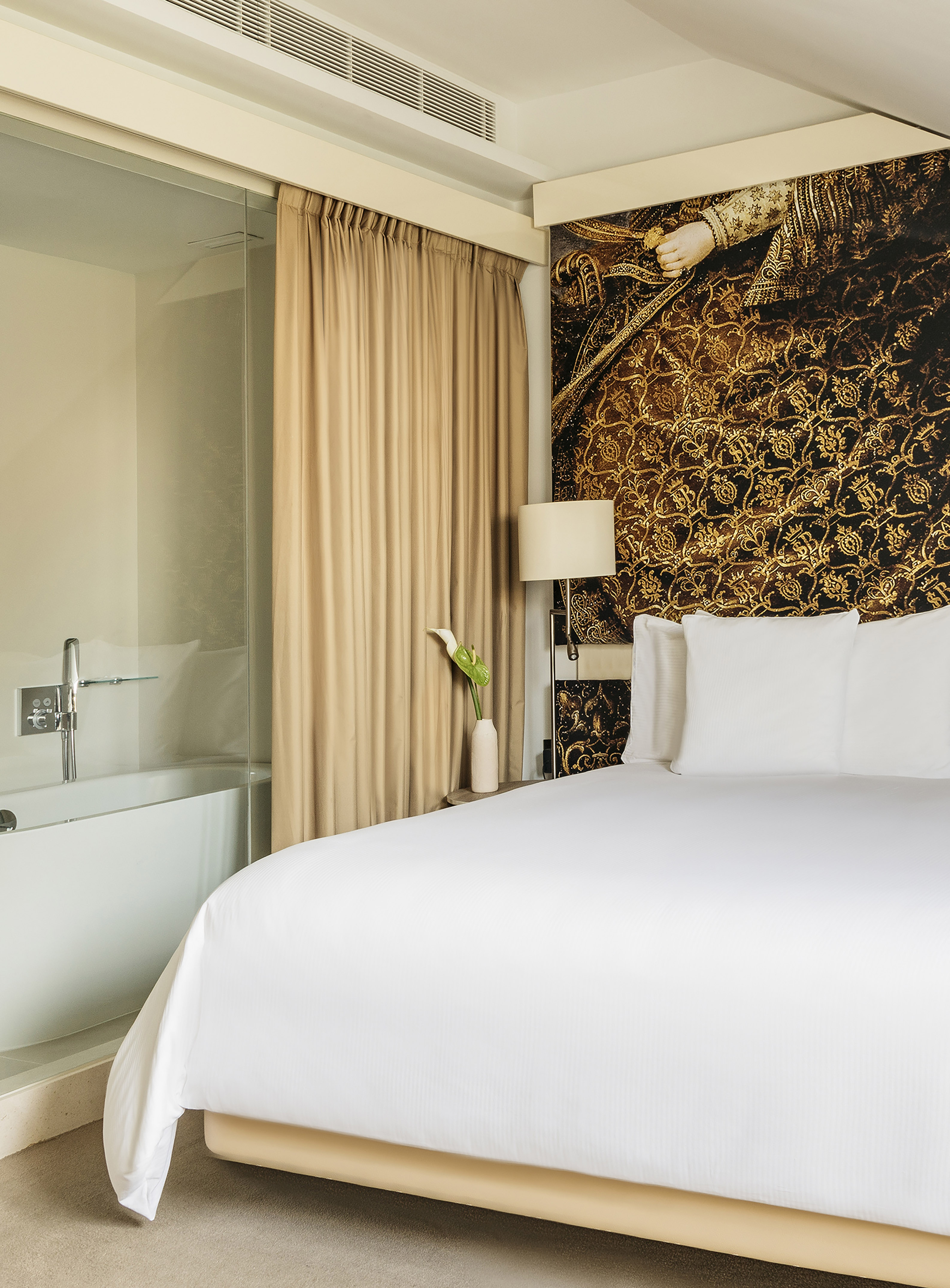 Modern hotel room with large bed, gold patterned wall, and open-concept bathtub.
