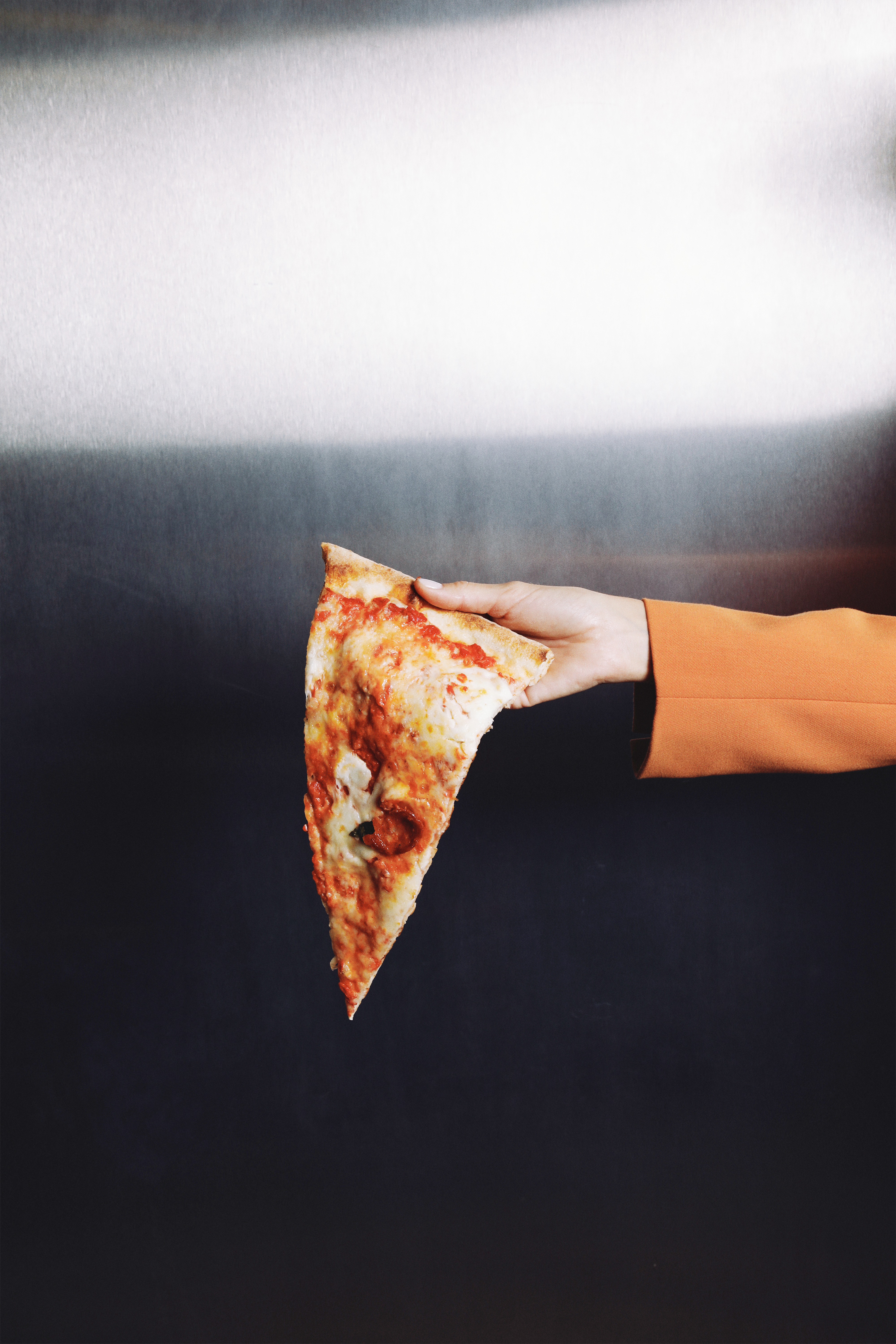 Hand in orange jacket holds a bitten pizza slice with cheese and sauce.