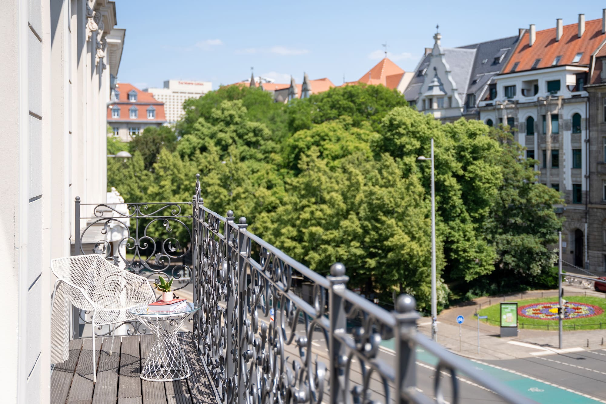 Apartment balcony with white chair, city view, green trees, and European architecture.