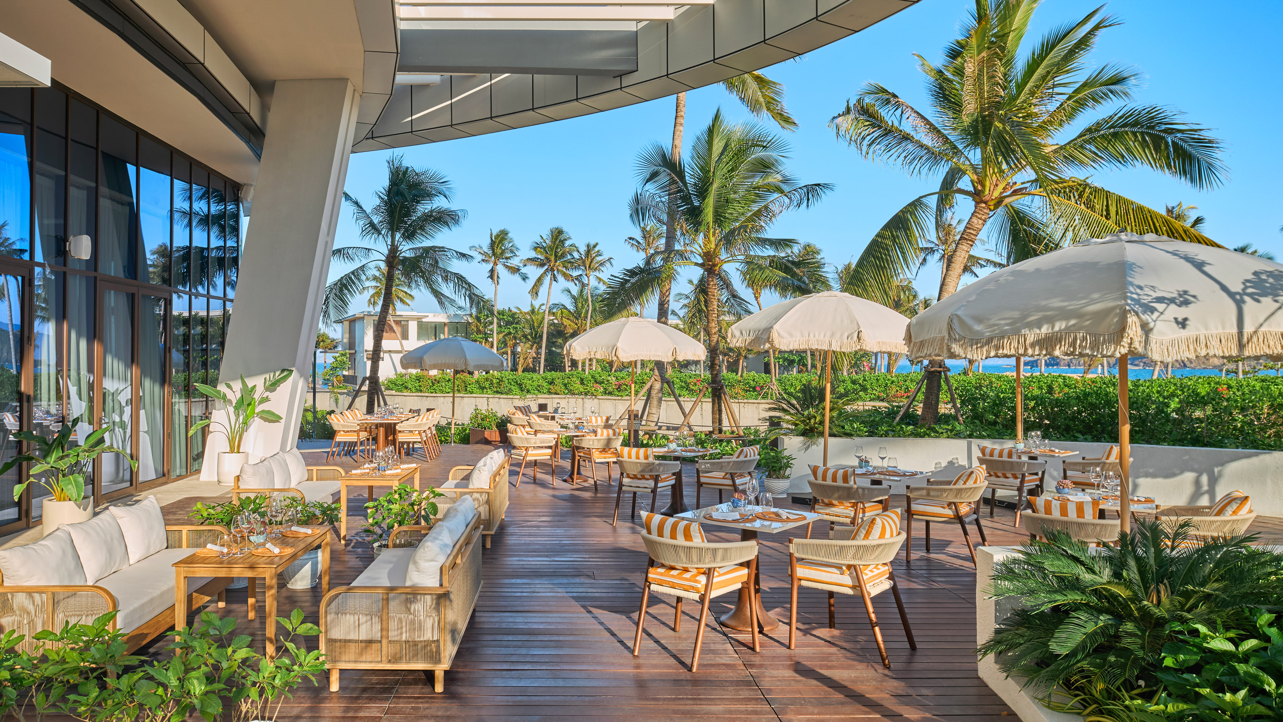 Beachfront restaurant terrace with dining tables, palm trees, and umbrellas.