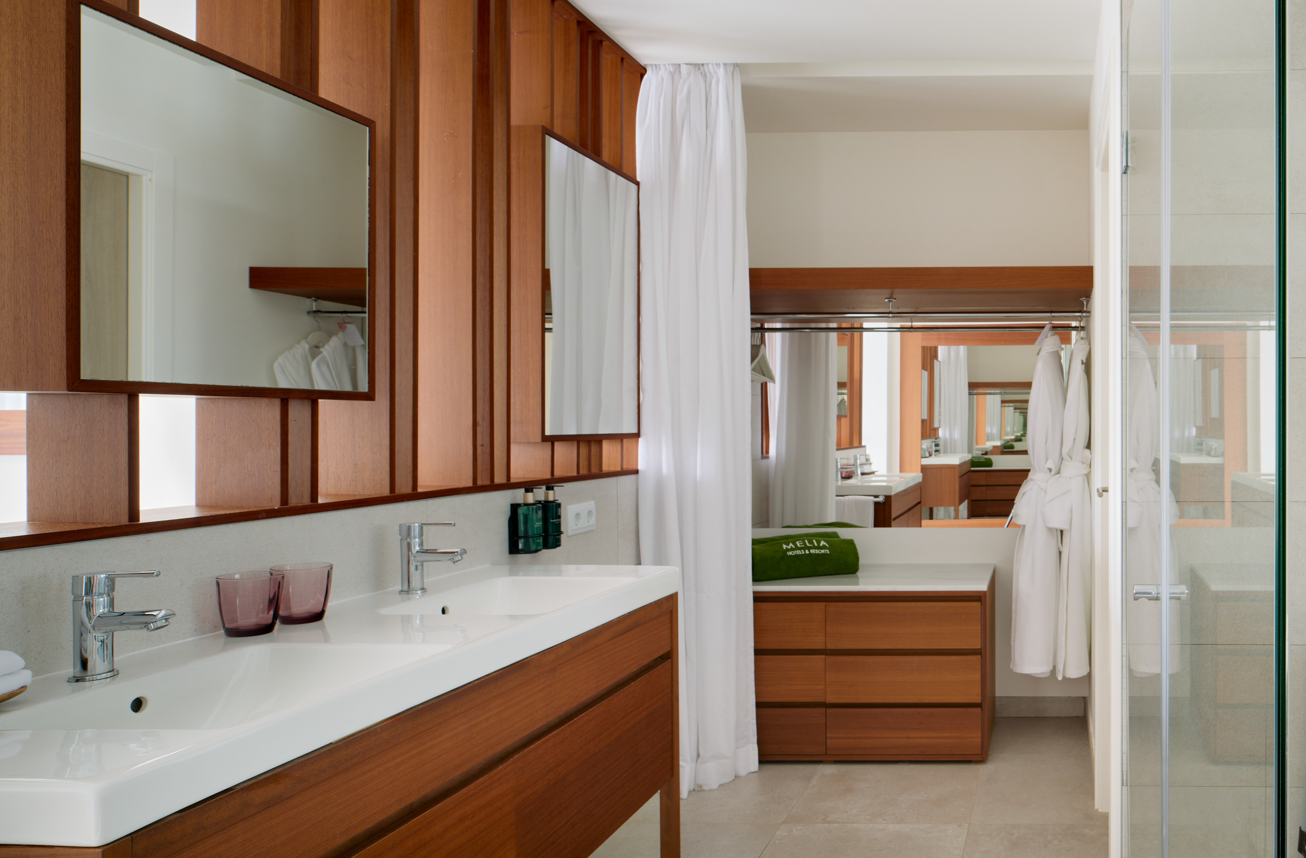 Wood-paneled hotel bathroom with double vanity, mirrors, bathrobes, and shower cabin.