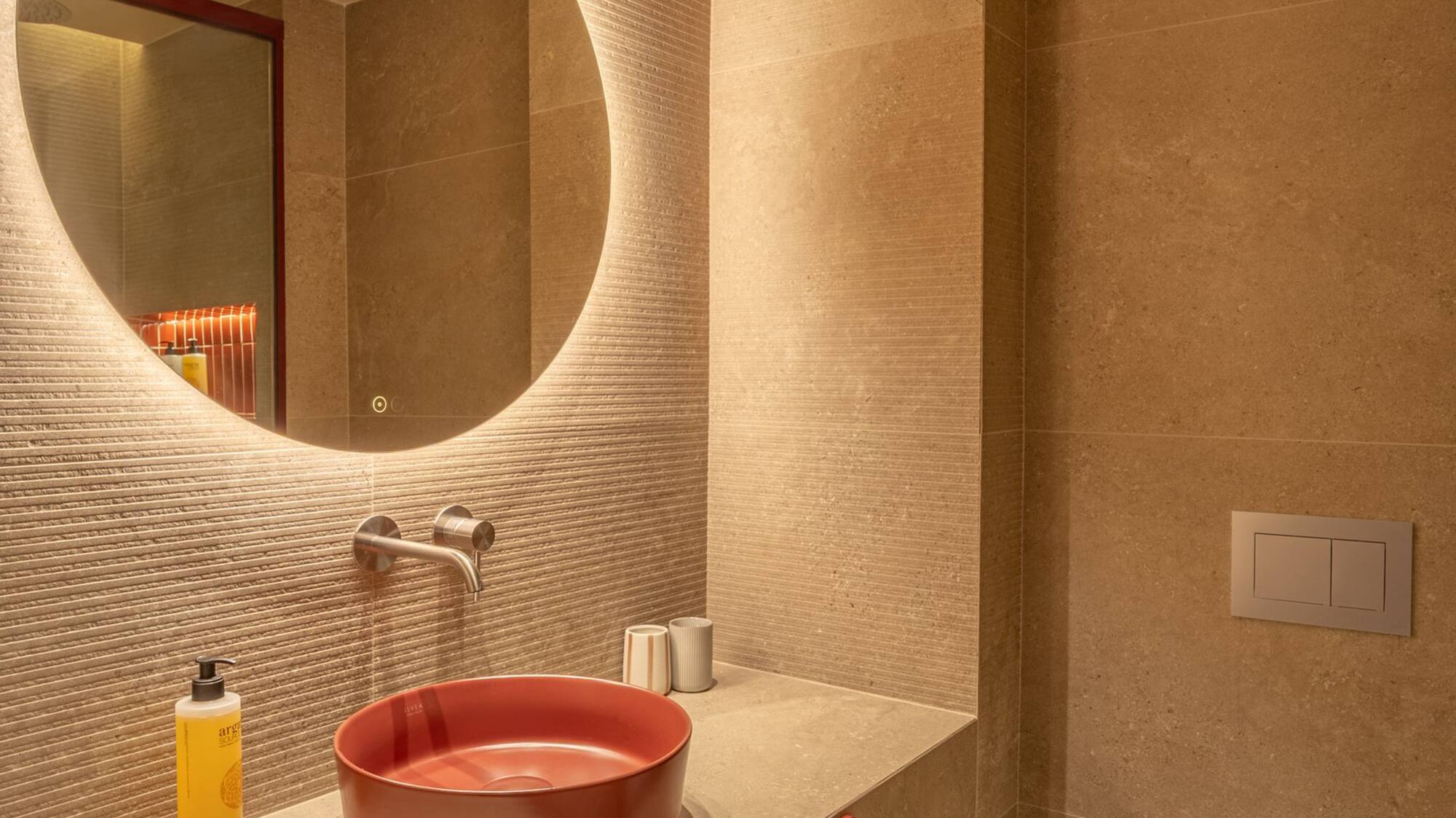 Modern hotel bathroom with red vessel sink, backlit round mirror, beige tiles.