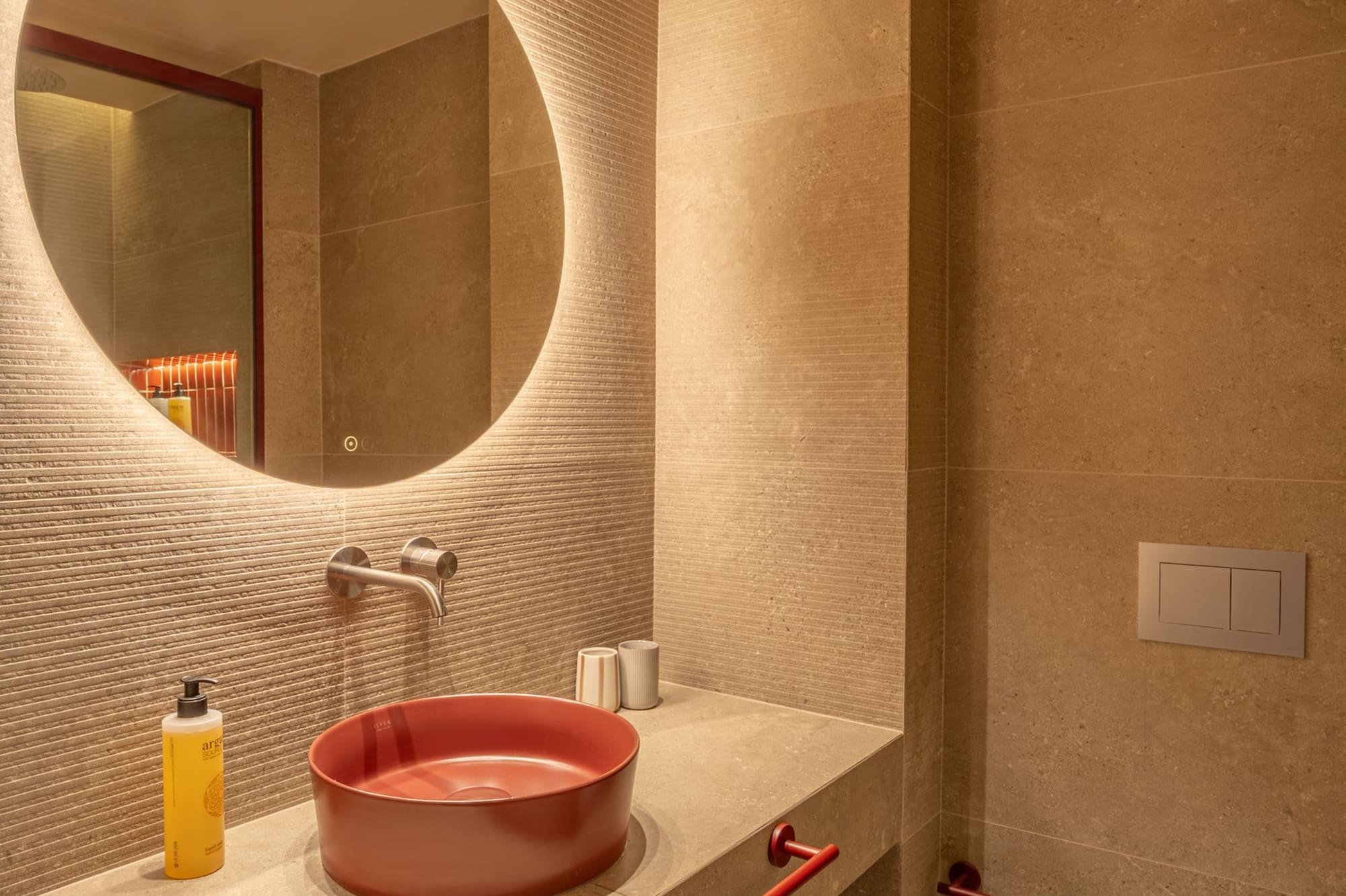 Modern hotel bathroom with red vessel sink, backlit round mirror, beige tiles.