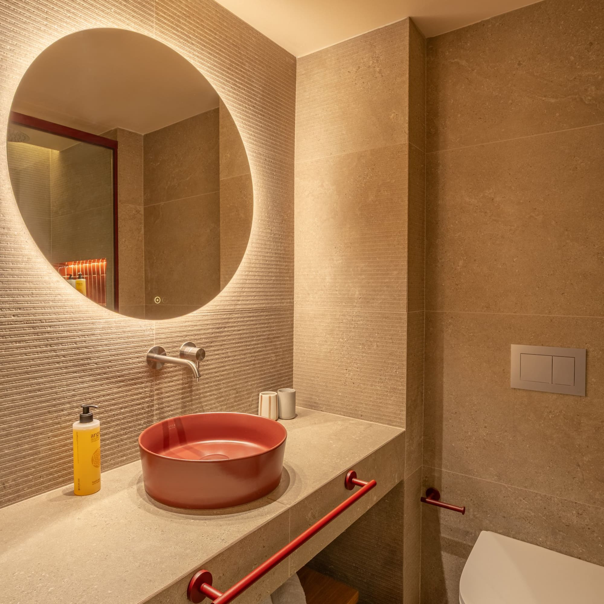 Modern hotel bathroom with red vessel sink, backlit round mirror, beige tiles.