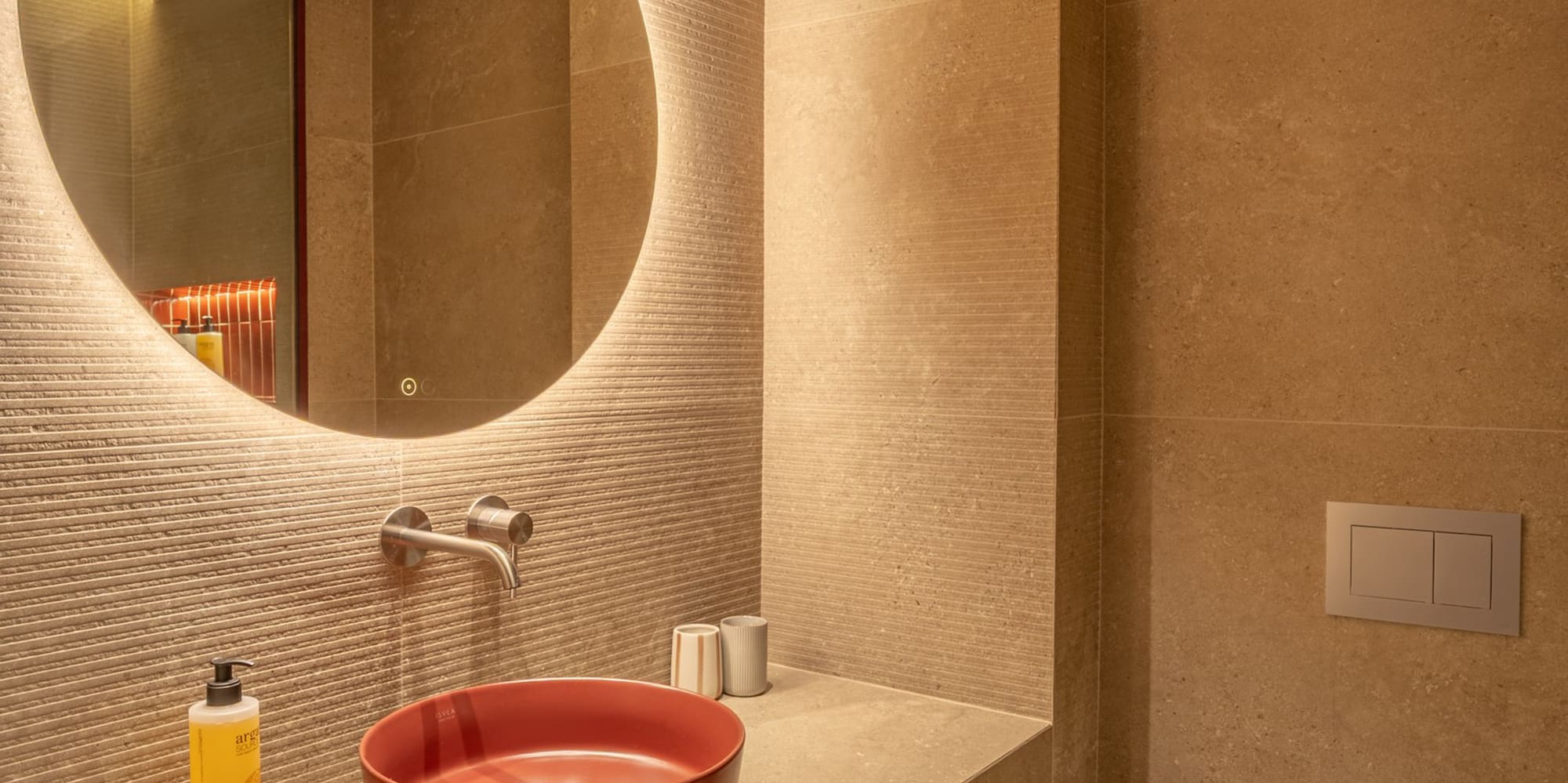 Modern hotel bathroom with red vessel sink, backlit round mirror, beige tiles.