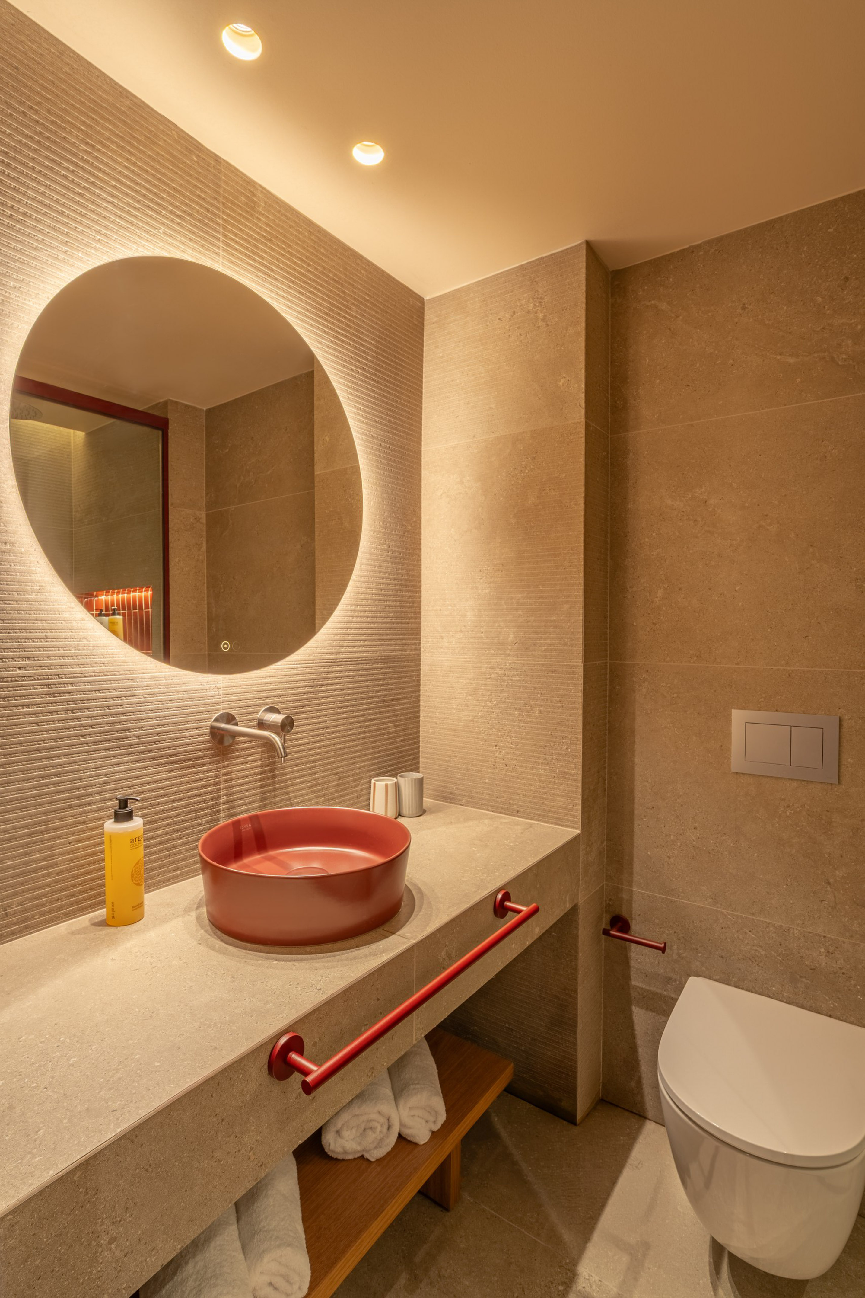 Modern hotel bathroom with red vessel sink, backlit round mirror, beige tiles.