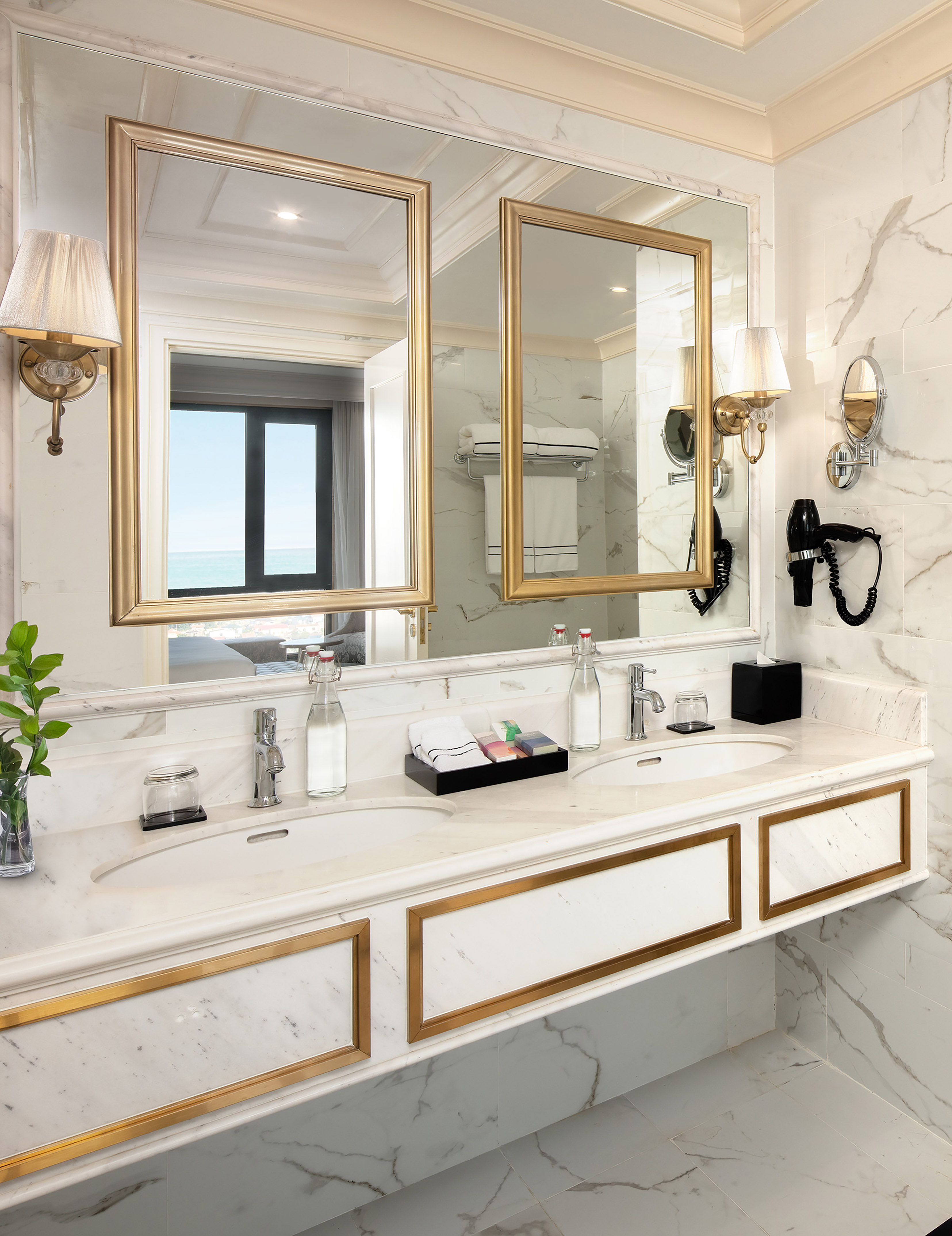 Luxury hotel bathroom with dual marble vanity, gold mirrors, and ocean view reflection.