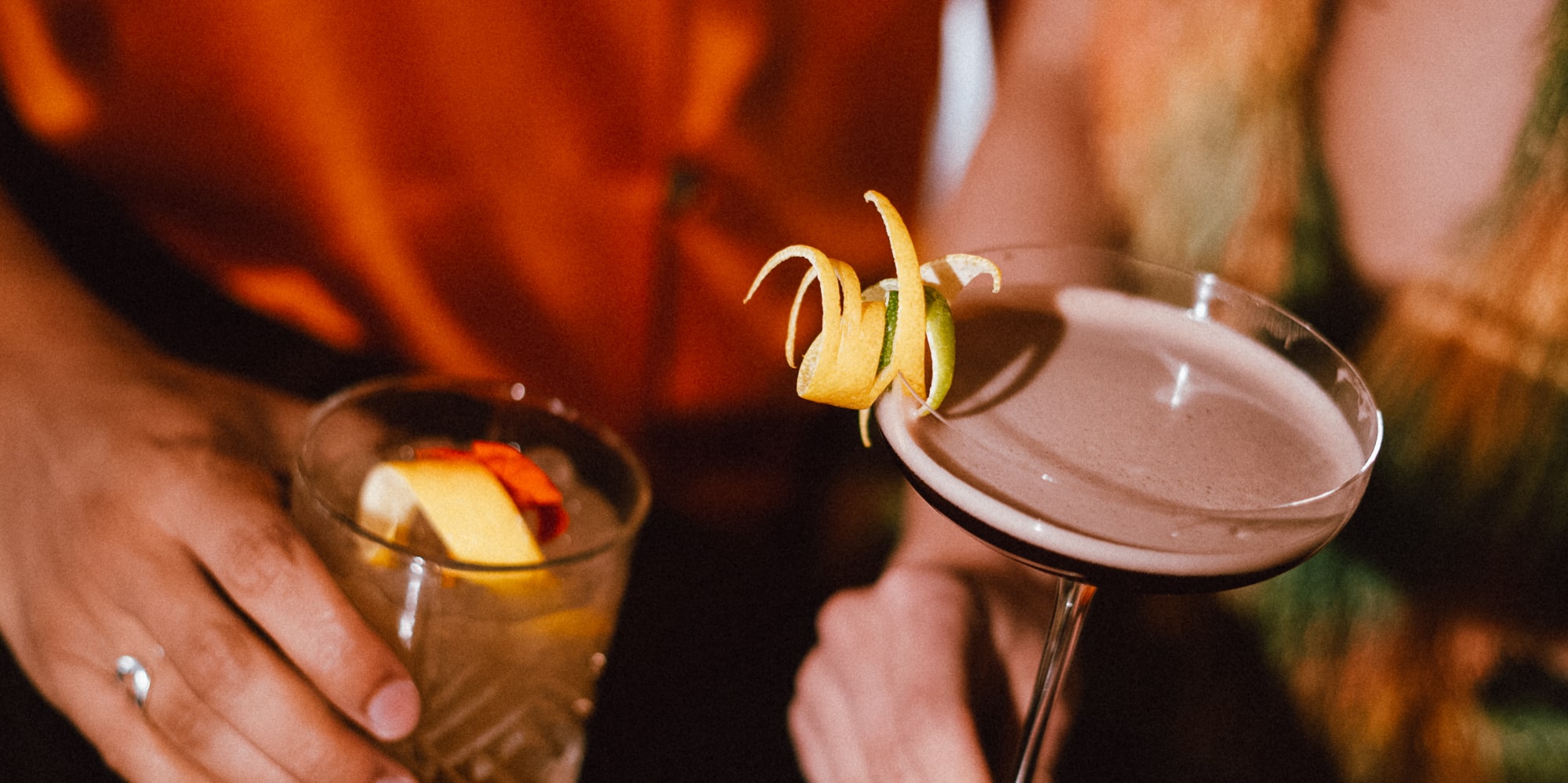 Two hands holding cocktails on a dark bar. Martini with citrus peel, old fashioned with lemon.