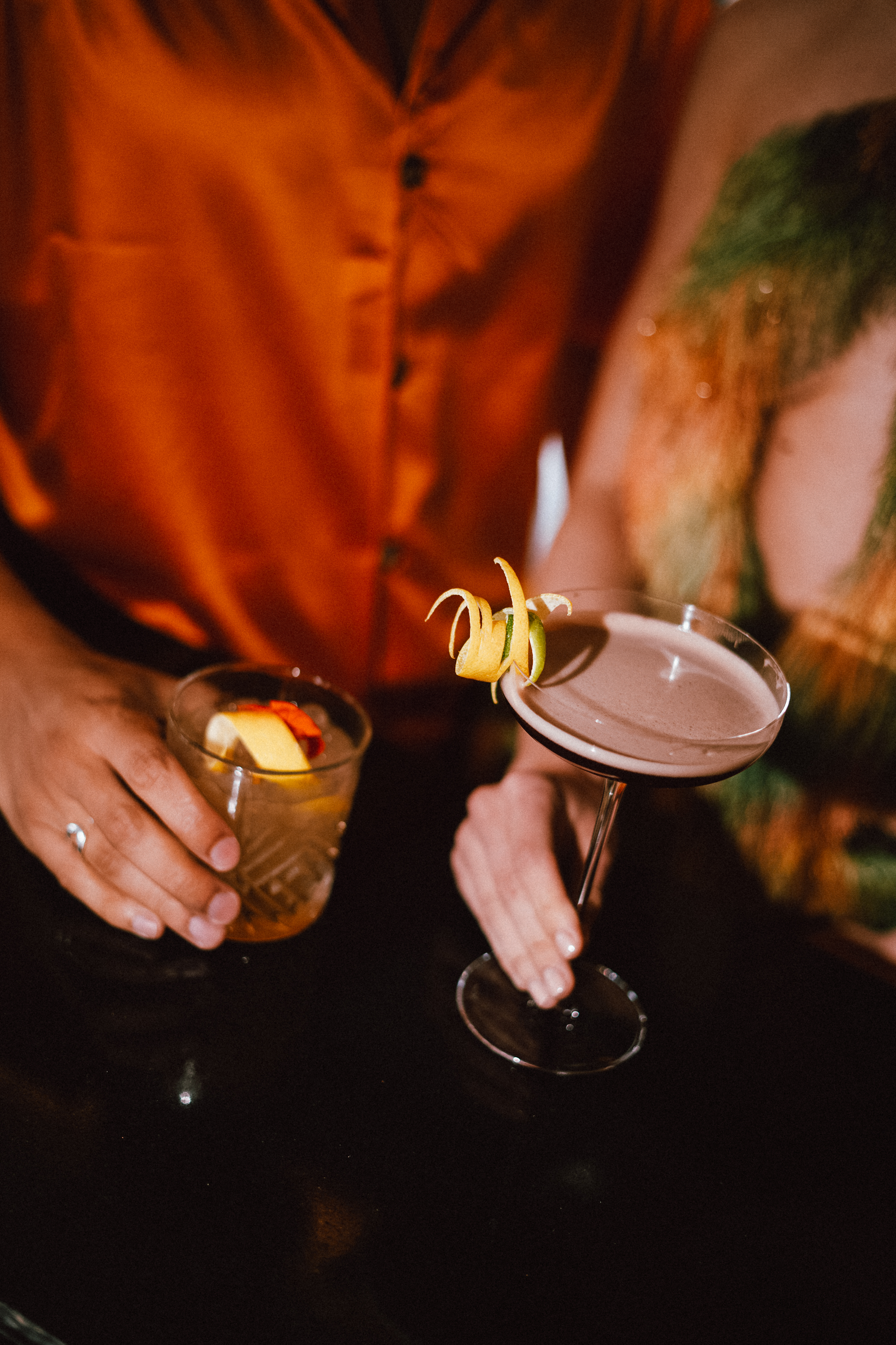 Two hands holding cocktails on a dark bar. Martini with citrus peel, old fashioned with lemon.
