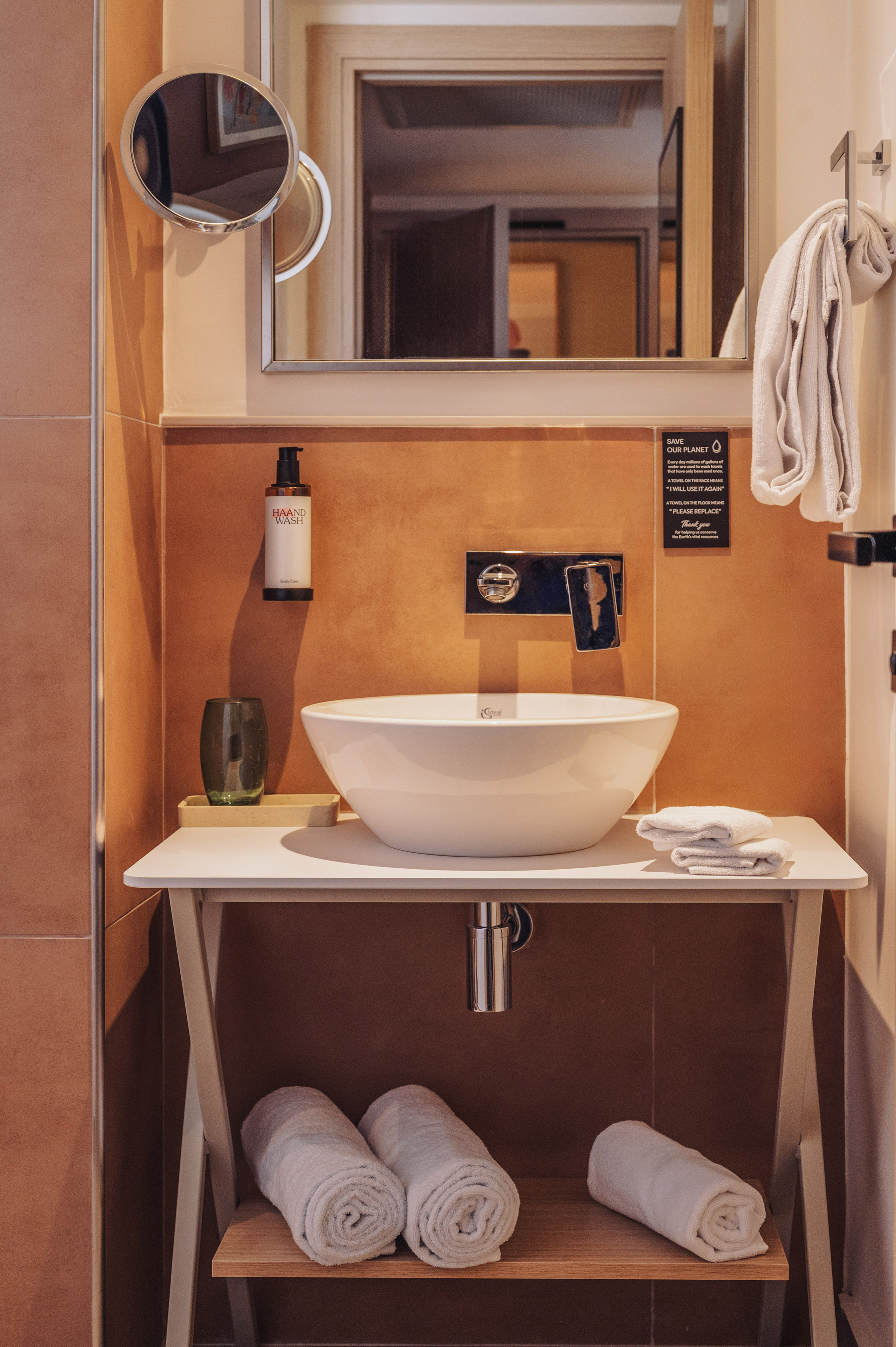 Modern bathroom sink area with hand wash, towels, mirror, and towel reuse sign.