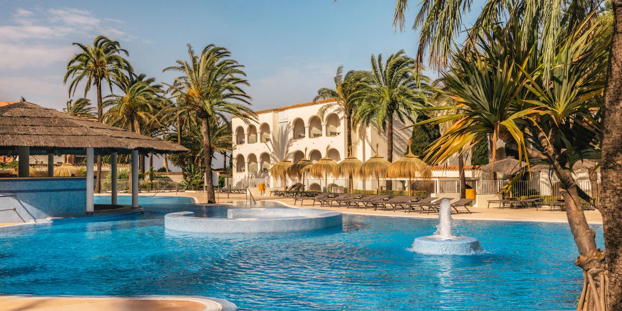 Tropical resort swimming pool with hotel, palm trees, loungers, thatched bar.