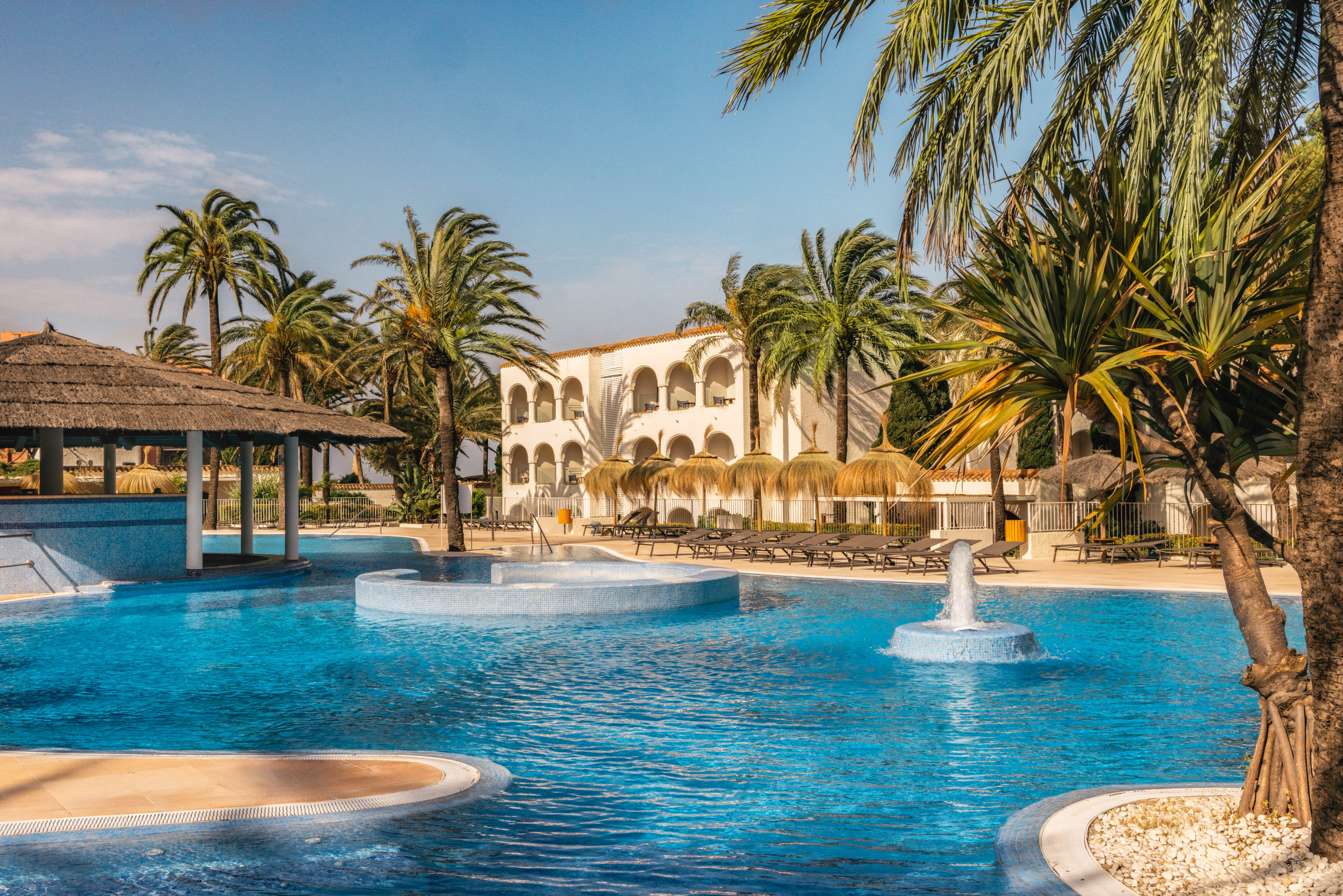 Tropical resort swimming pool with hotel, palm trees, loungers, thatched bar.