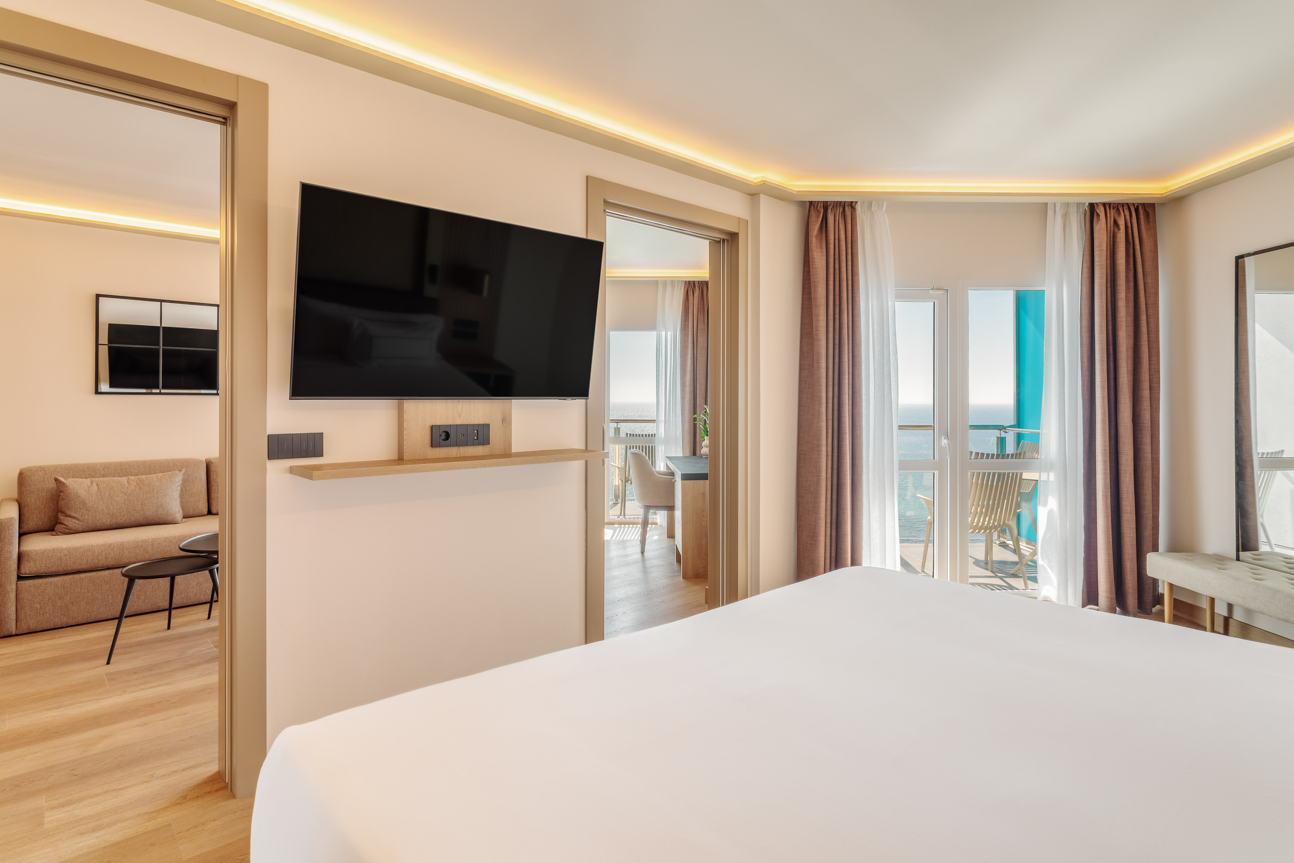 Modern hotel suite with ocean view balcony, large bed, TV, and living area.