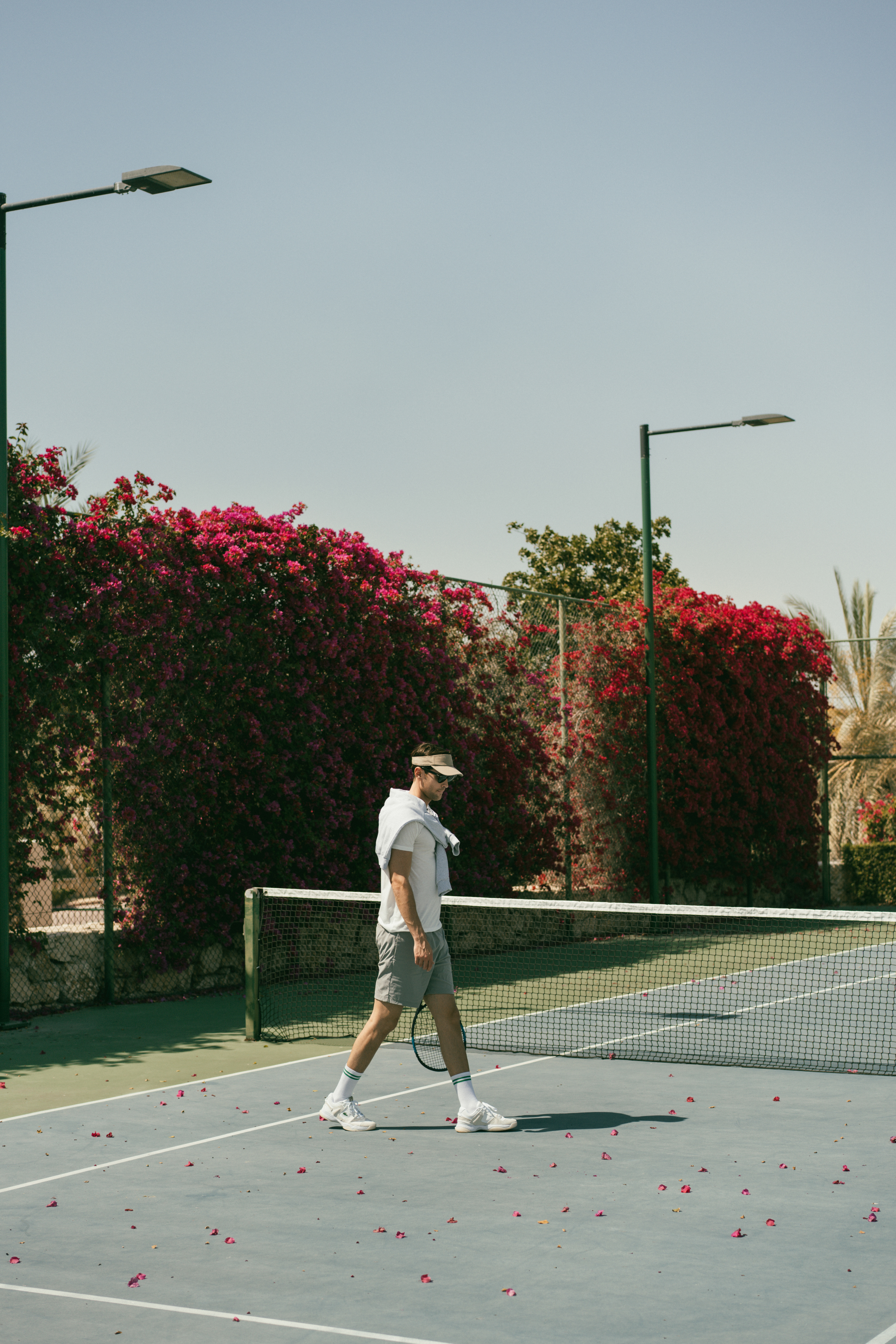 Tennis player walks on court with racket, pink petals, bougainvillea wall.