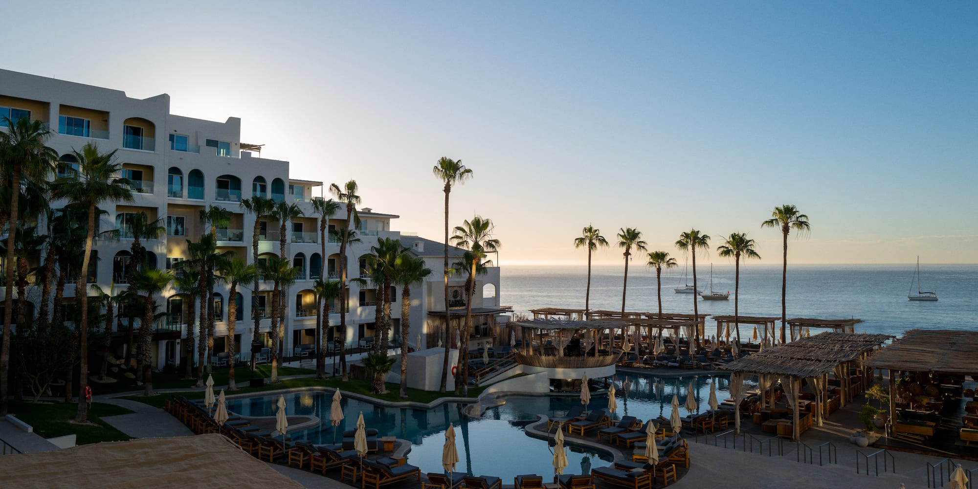 a pool with chairs and umbrellas next to a building with palm trees
