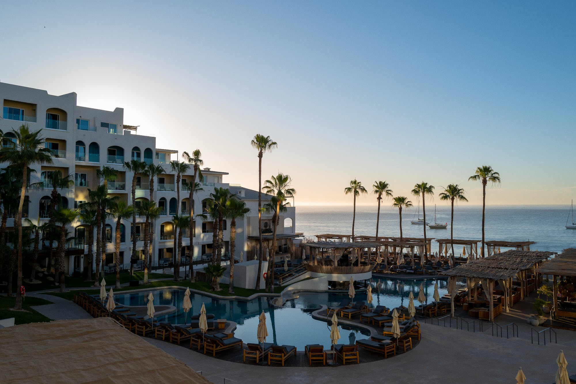 Oceanfront resort with pool, palm trees, and ocean view at sunset.