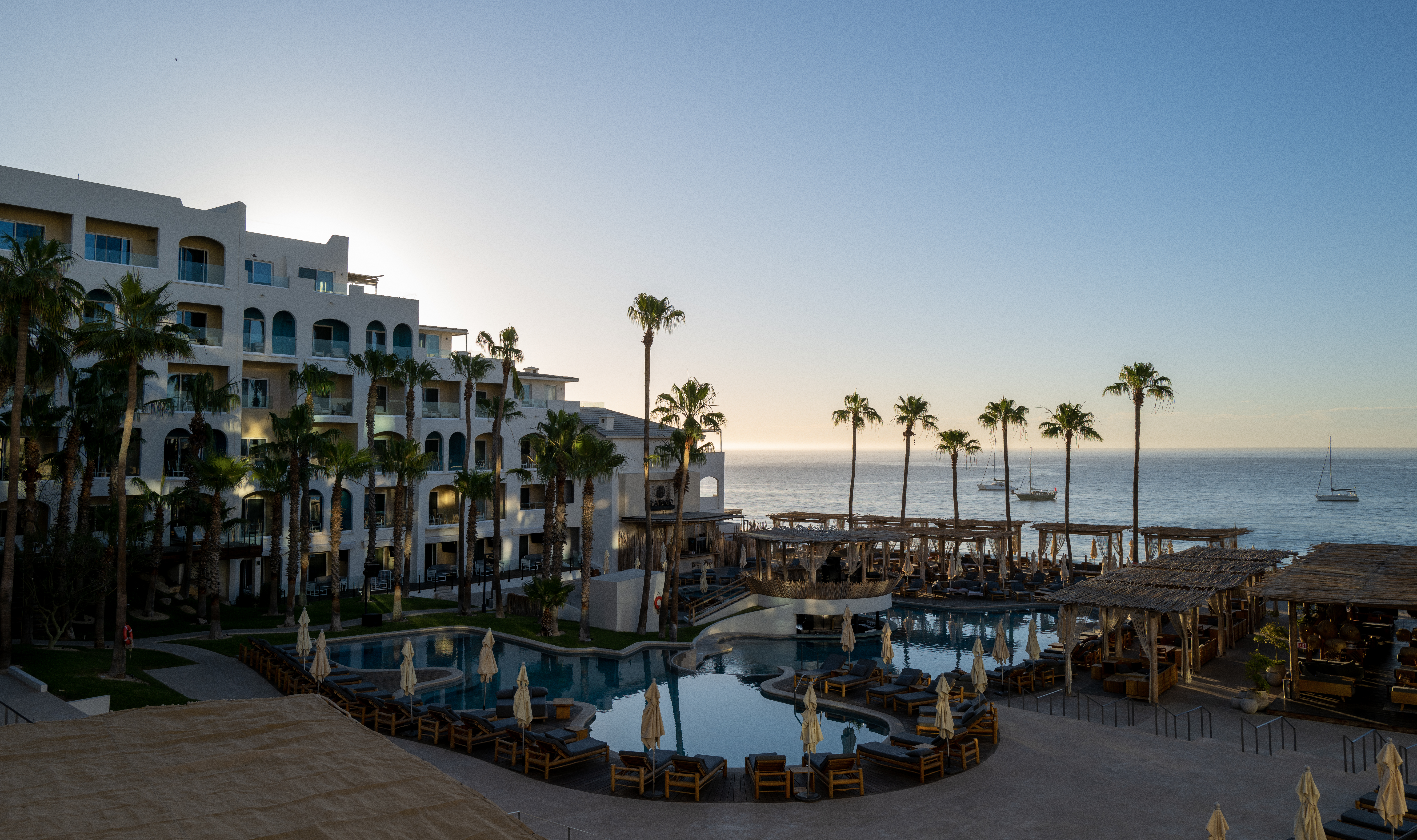 a pool with chairs and umbrellas next to a building with palm trees