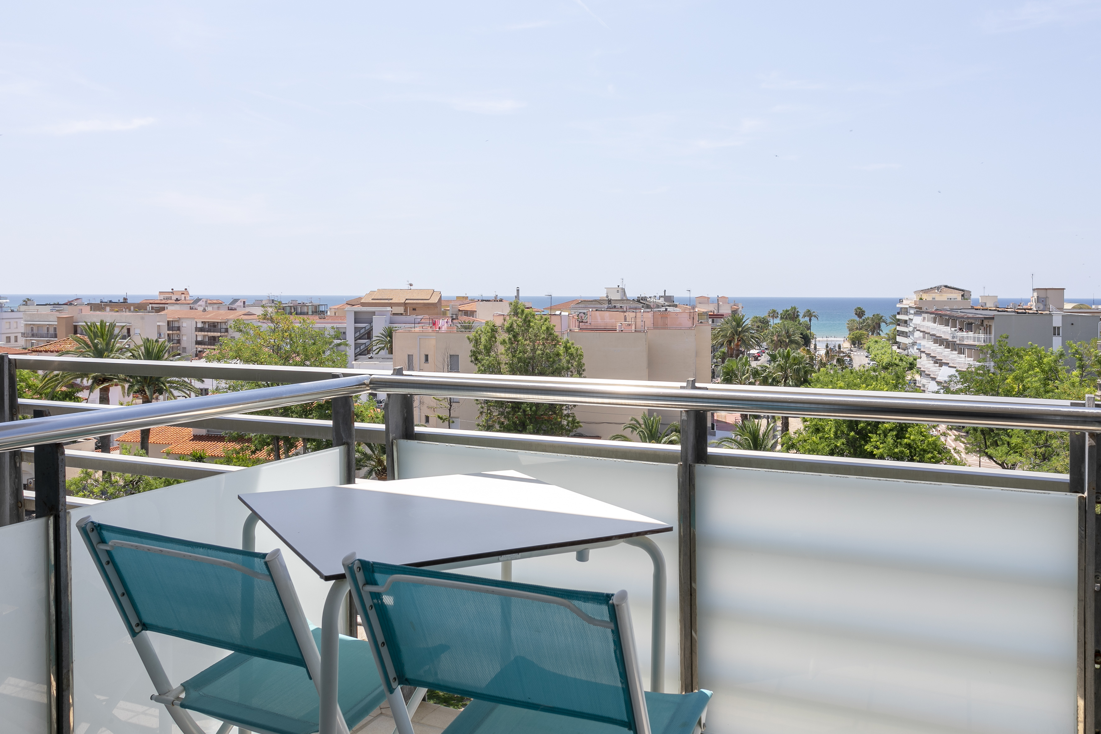 Balcony with turquoise chairs, table, and sea view of a sunny coastal town.