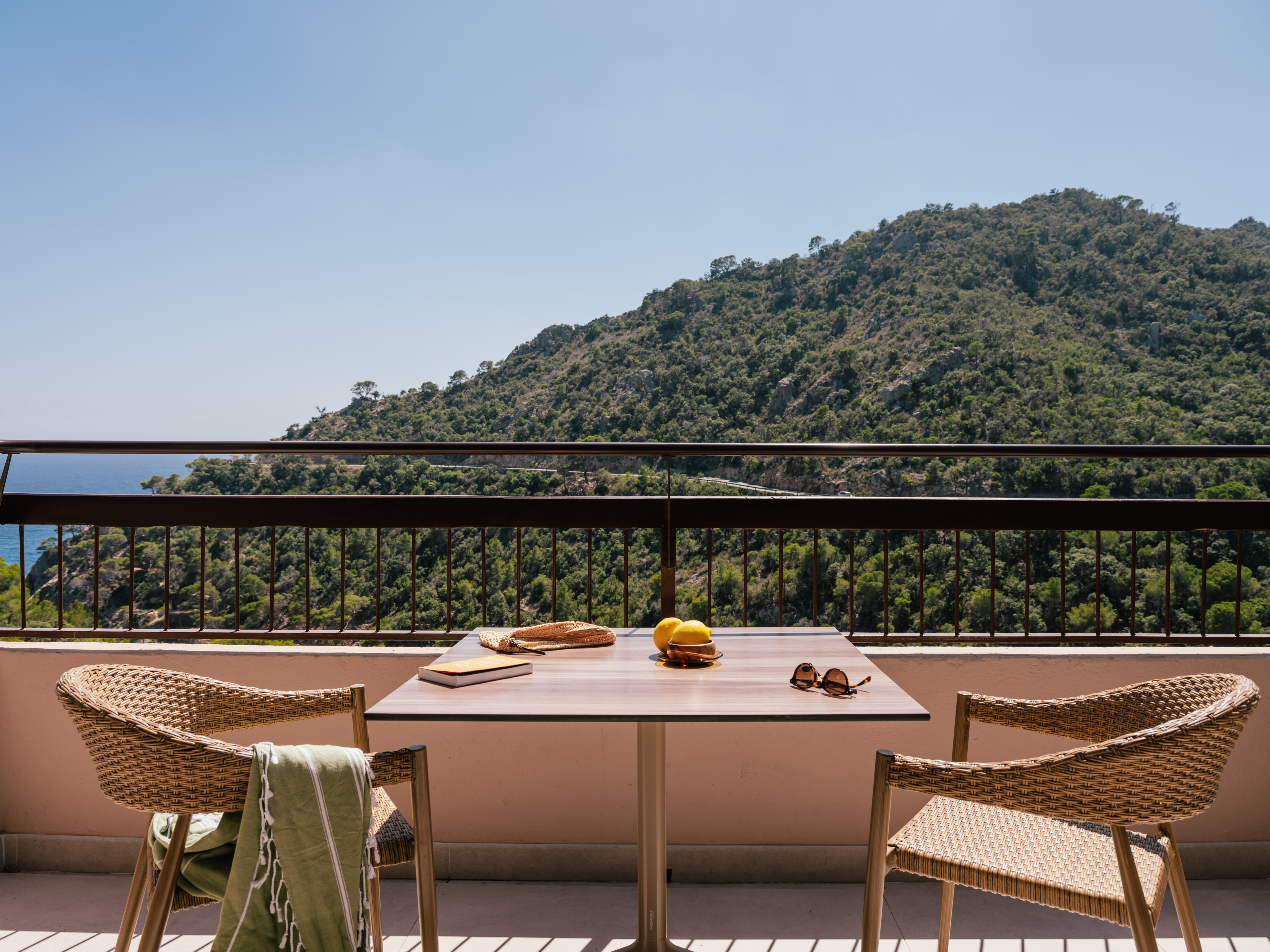 Balcony with rattan chairs, table, book, lemons, and scenic mountain-sea view.