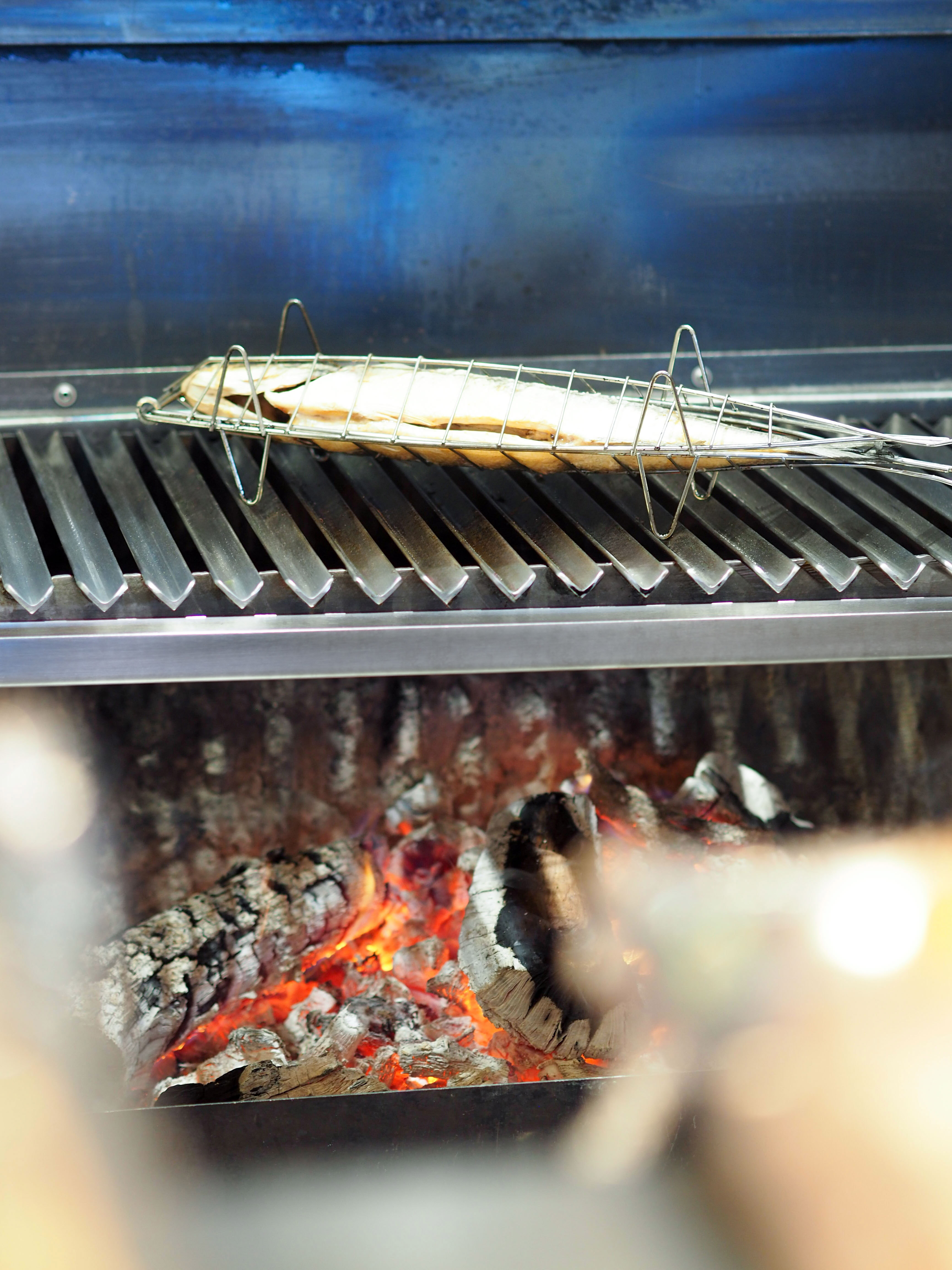 Whole fish in grilling basket over glowing wood-fired coals.