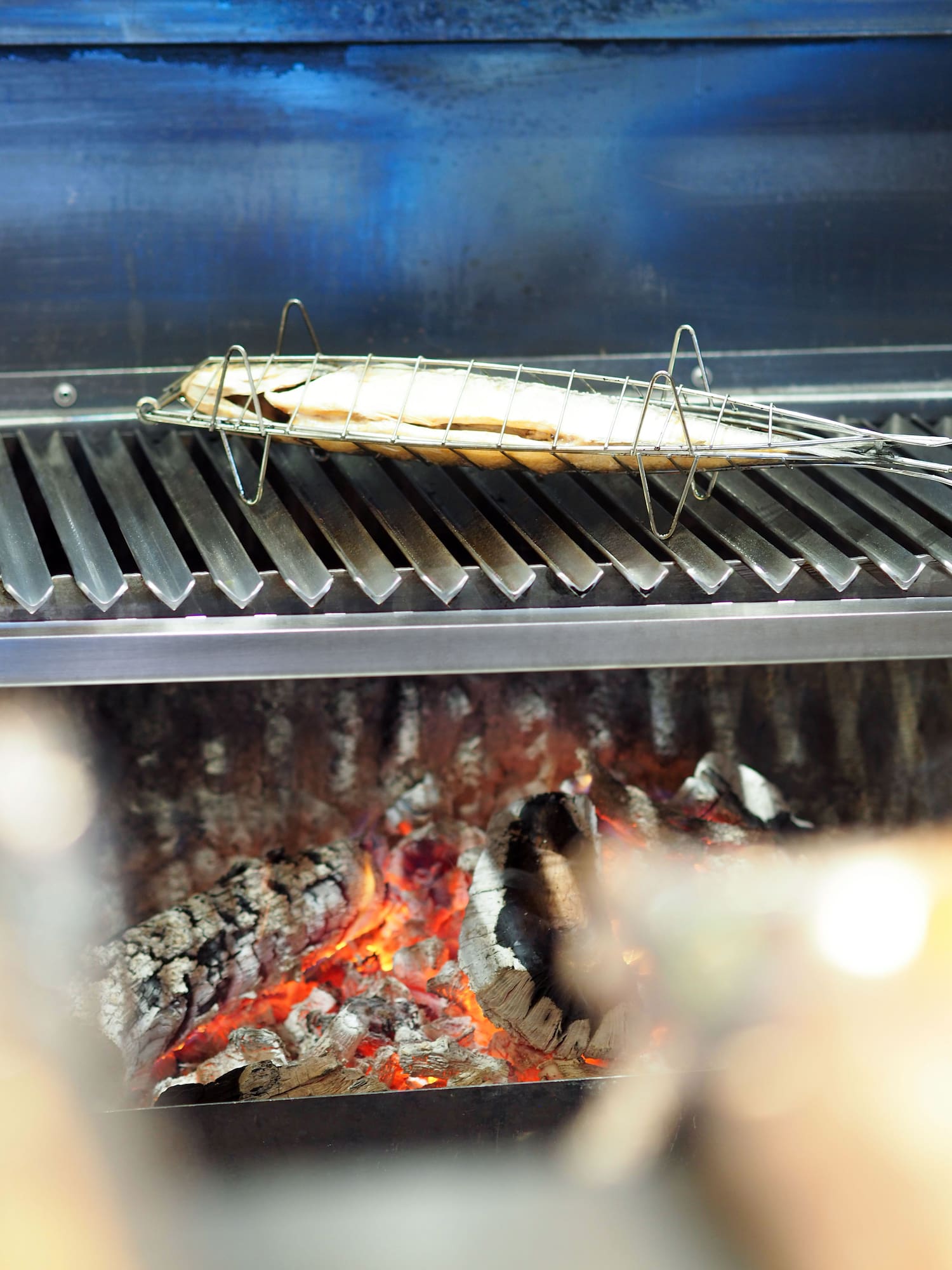 Whole fish in grilling basket over glowing wood-fired coals.