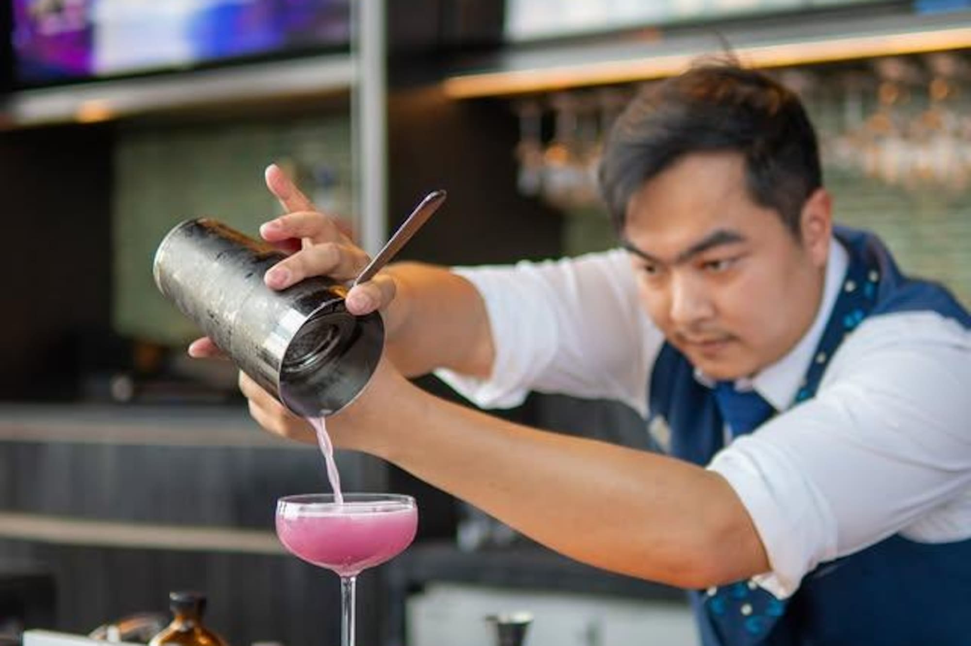 Mixologist pouring vibrant pink cocktail from shaker at a modern bar.