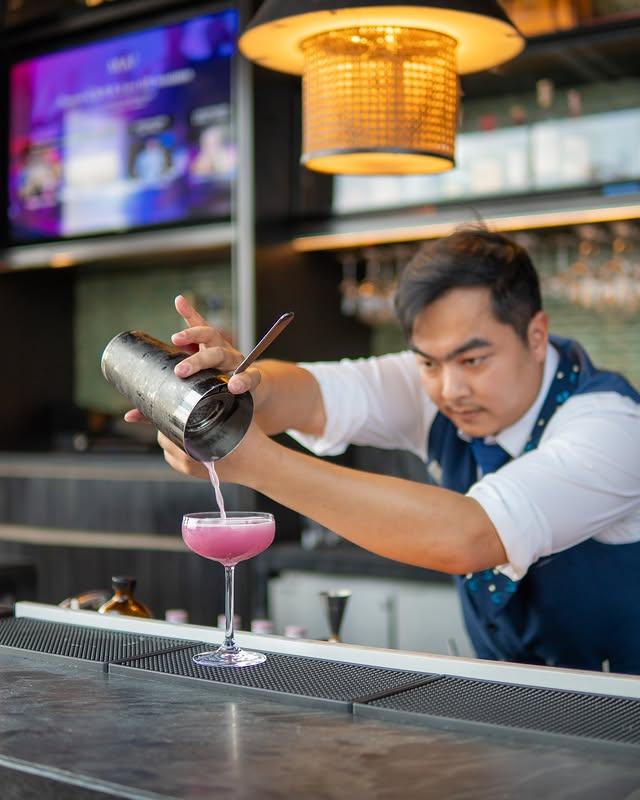 Mixologist pouring vibrant pink cocktail from shaker at a modern bar.