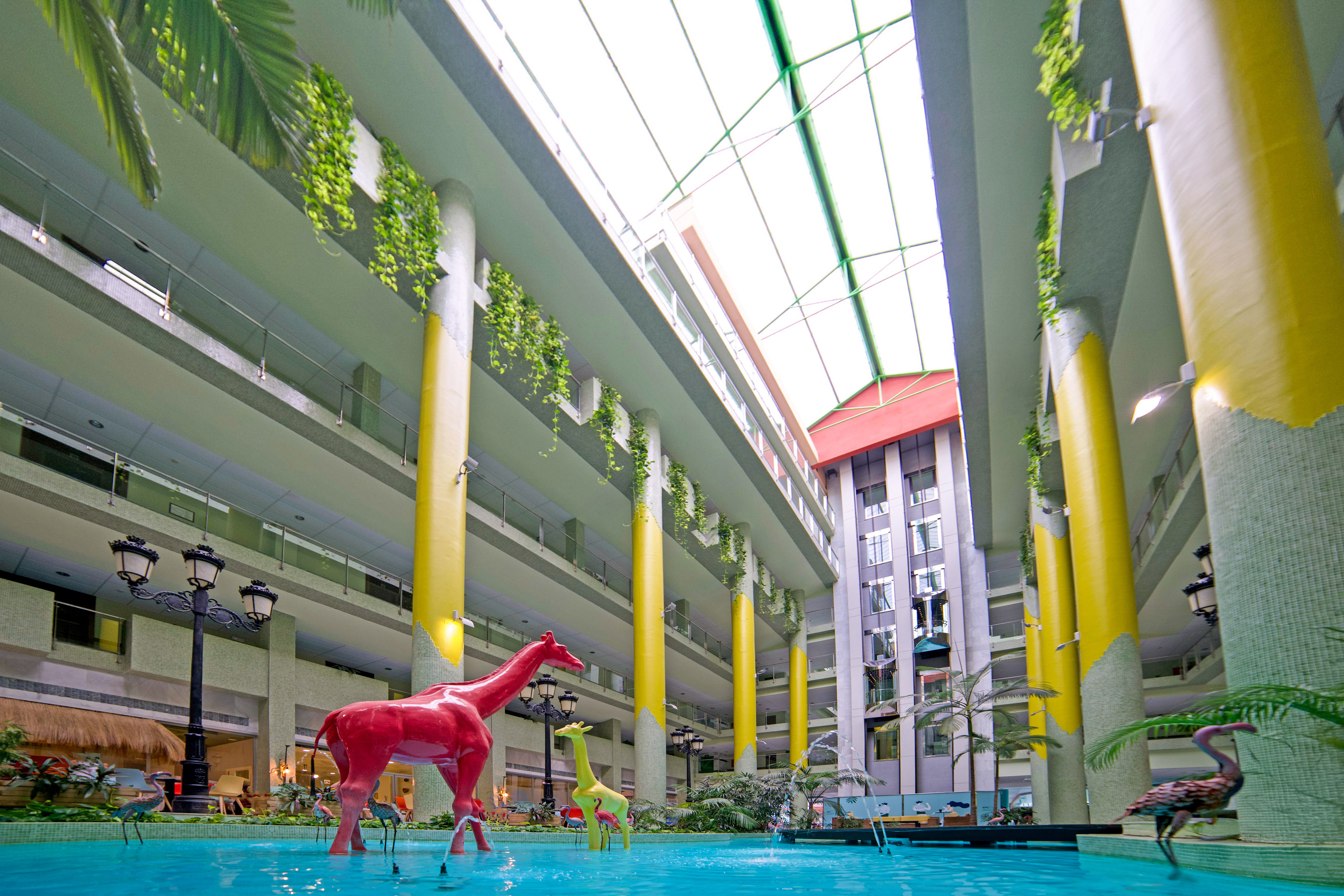 Red giraffe sculpture in a bright atrium with yellow pillars and a water feature.