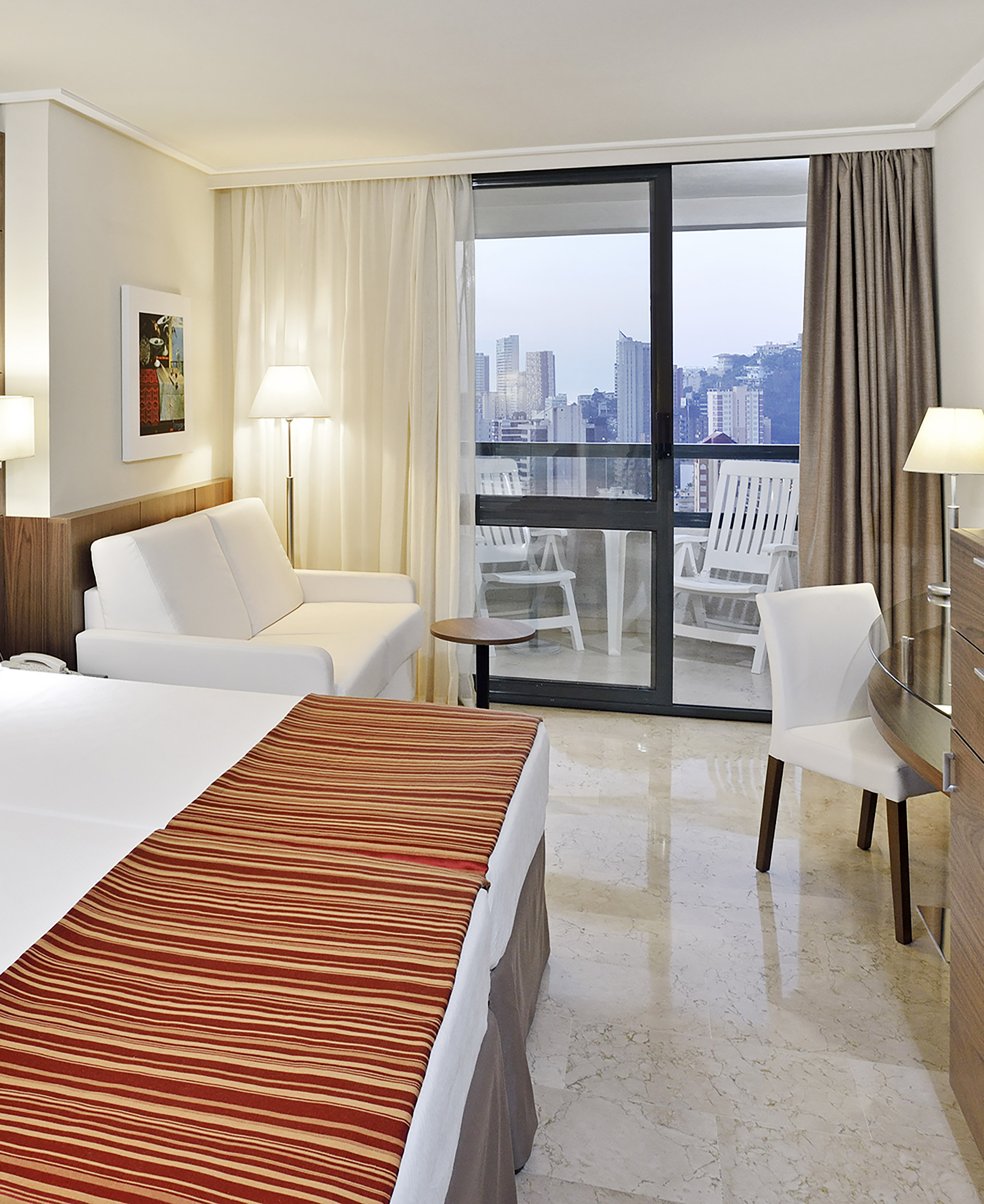 Modern hotel room with bed, sofa, work desk, and balcony city view.