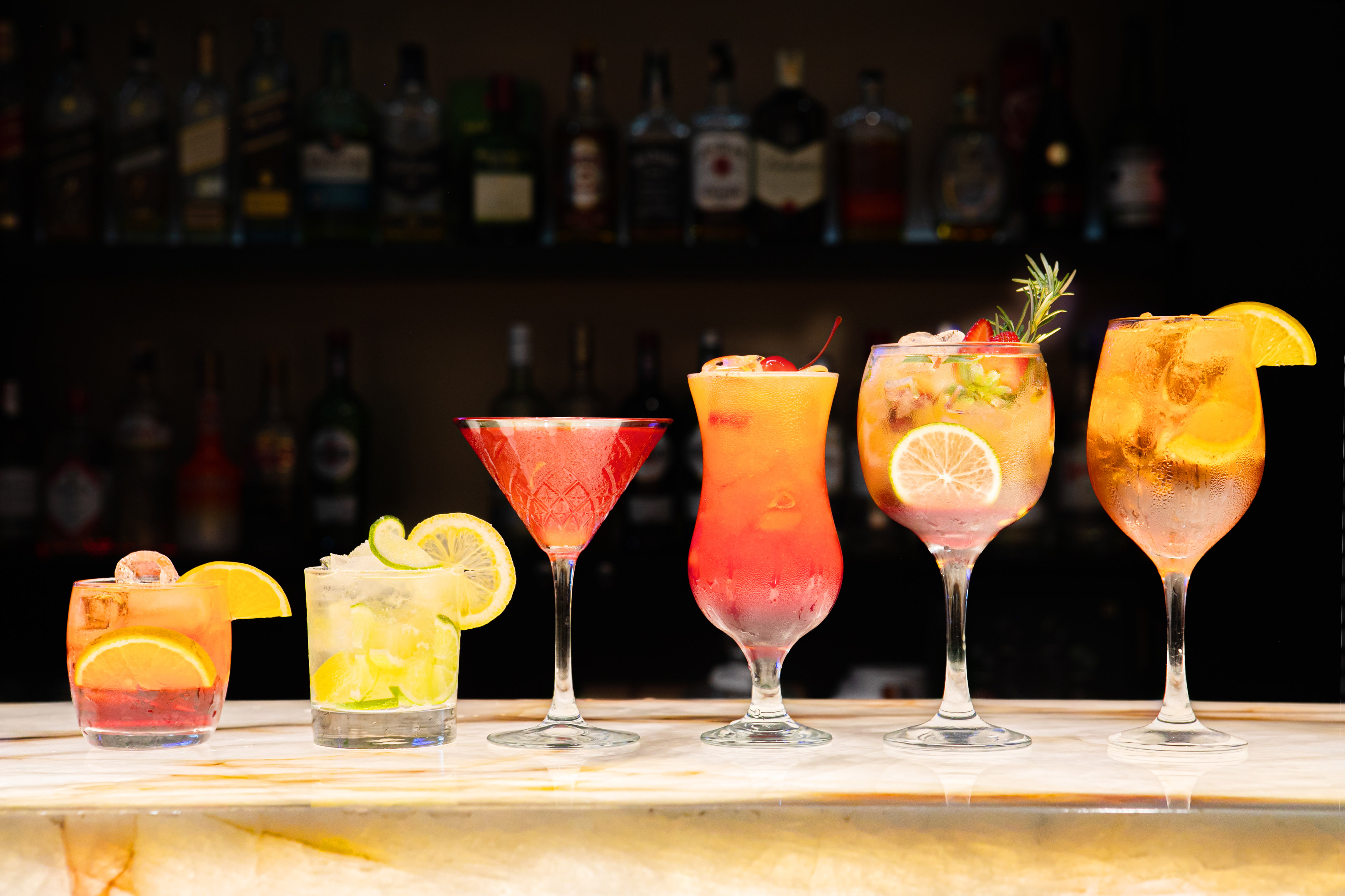 Six vibrant garnished cocktails on bar counter, blurred liquor bottles.