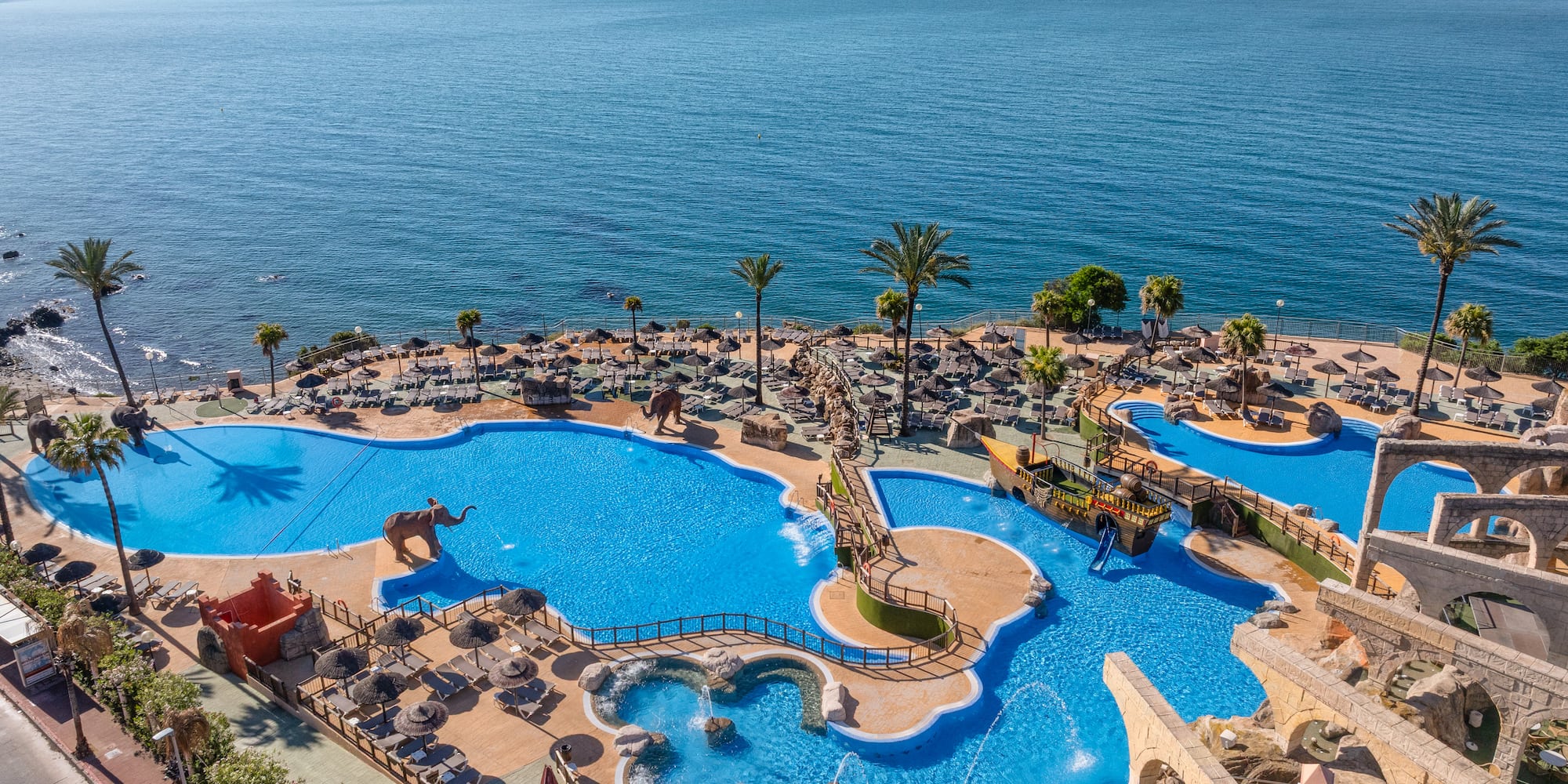 Seaside resort water park with themed pools, palm trees, and ocean views.