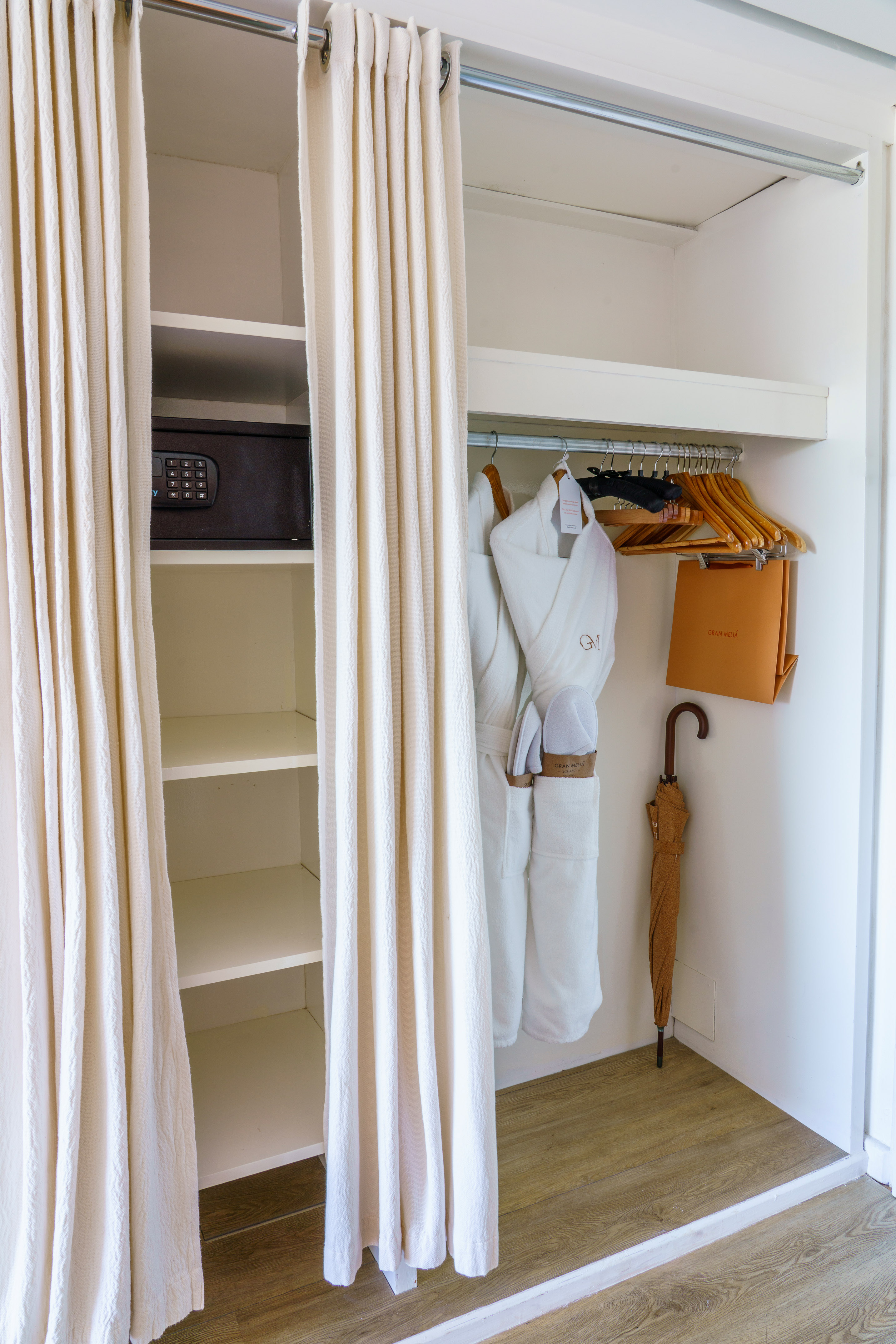 Hotel wardrobe with safe, bathrobes, slippers, umbrella, and cream curtains.
