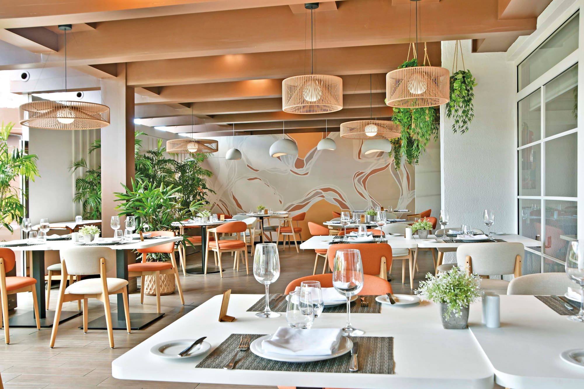 Modern restaurant interior with orange chairs, rattan lights, and lush plants.