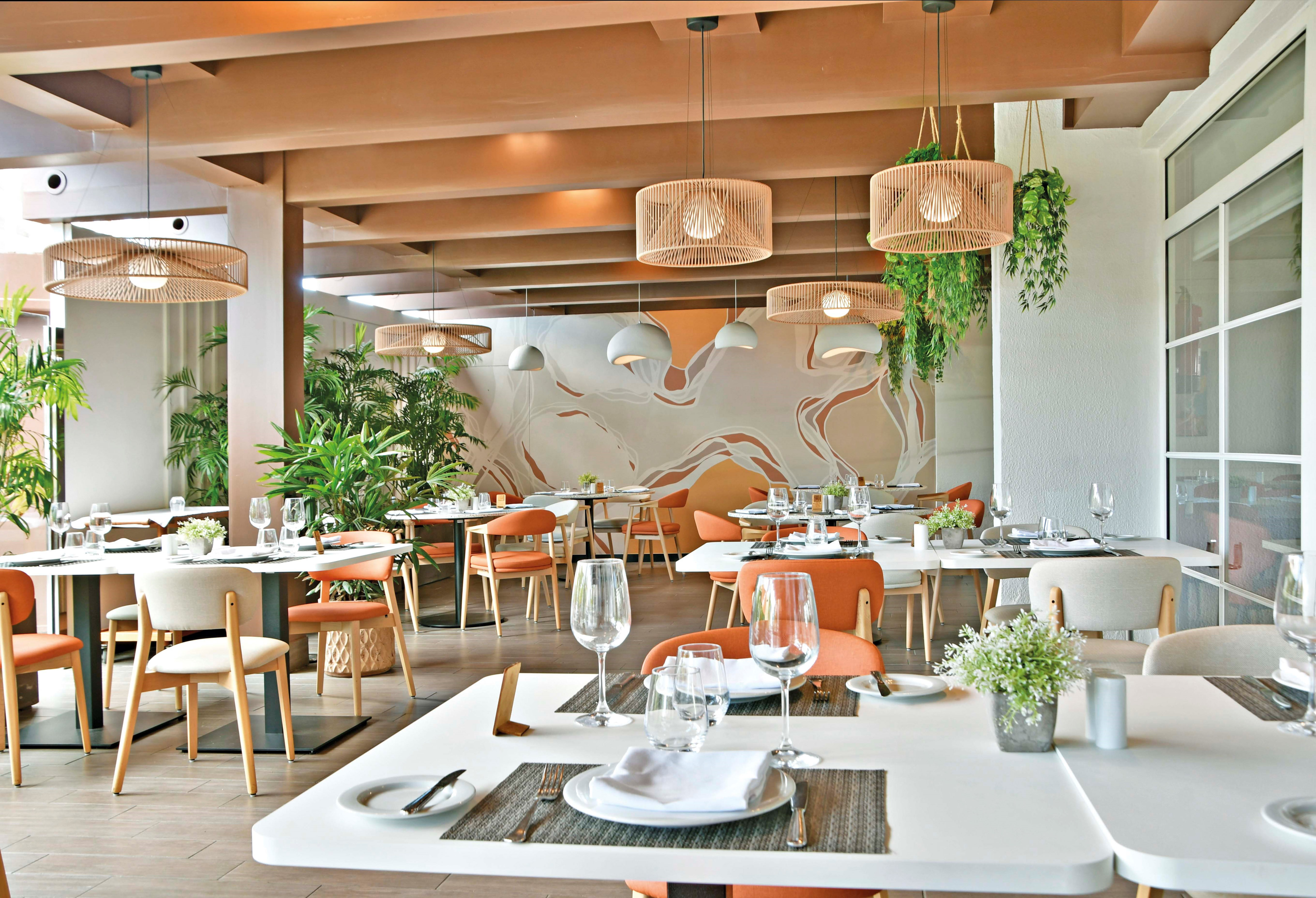 Modern restaurant interior with orange chairs, rattan lights, and lush plants.