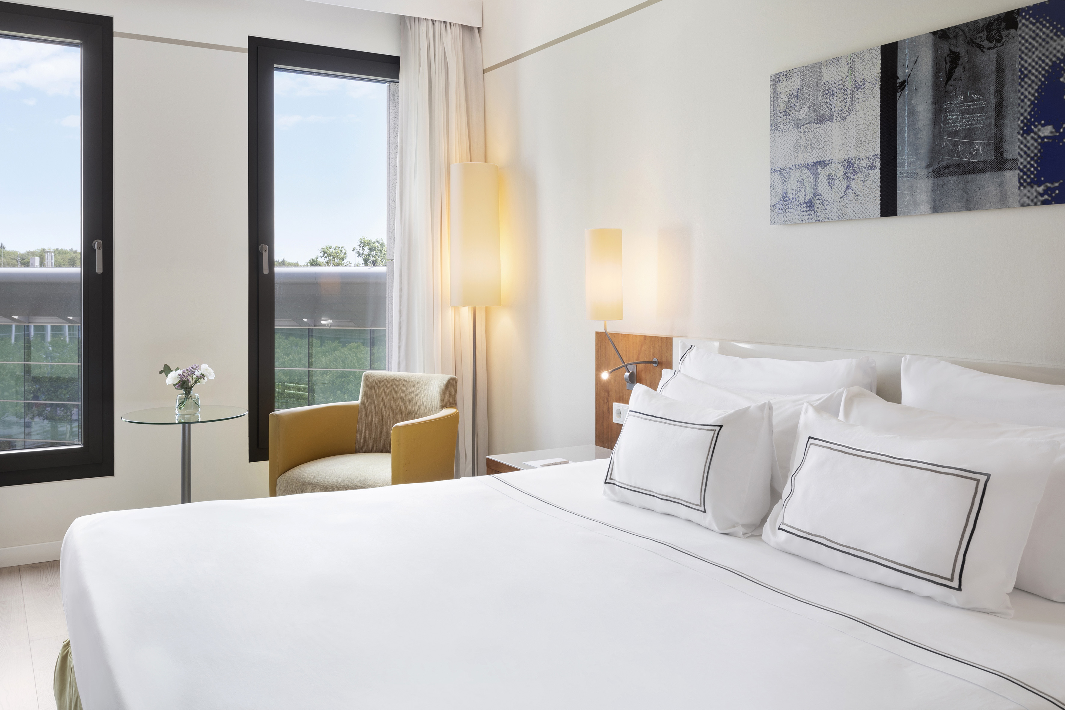 Modern hotel room with white bed, large windows, yellow armchair, and abstract art.