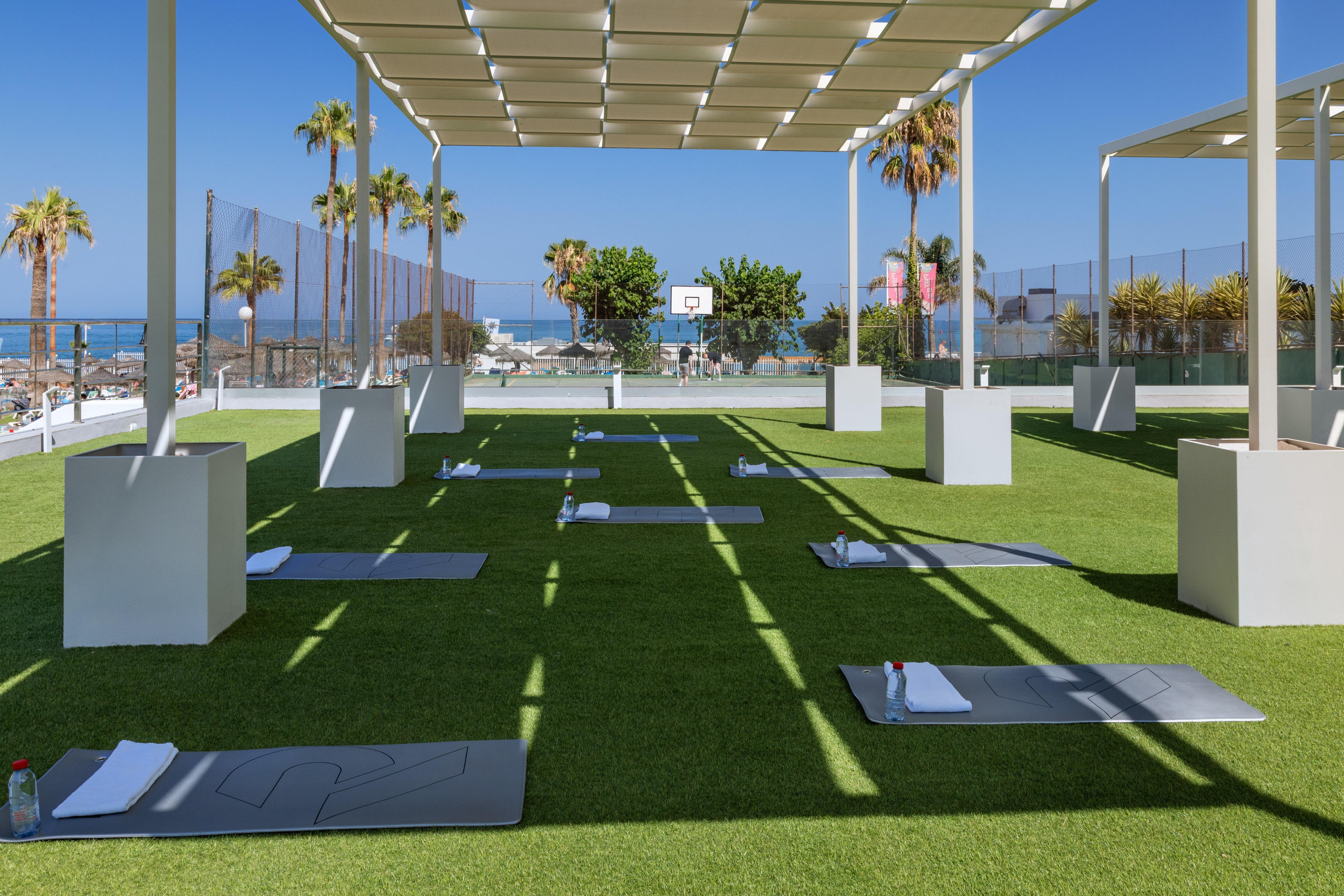 Outdoor yoga mats on green turf under pergola, overlooking beach and palm trees.