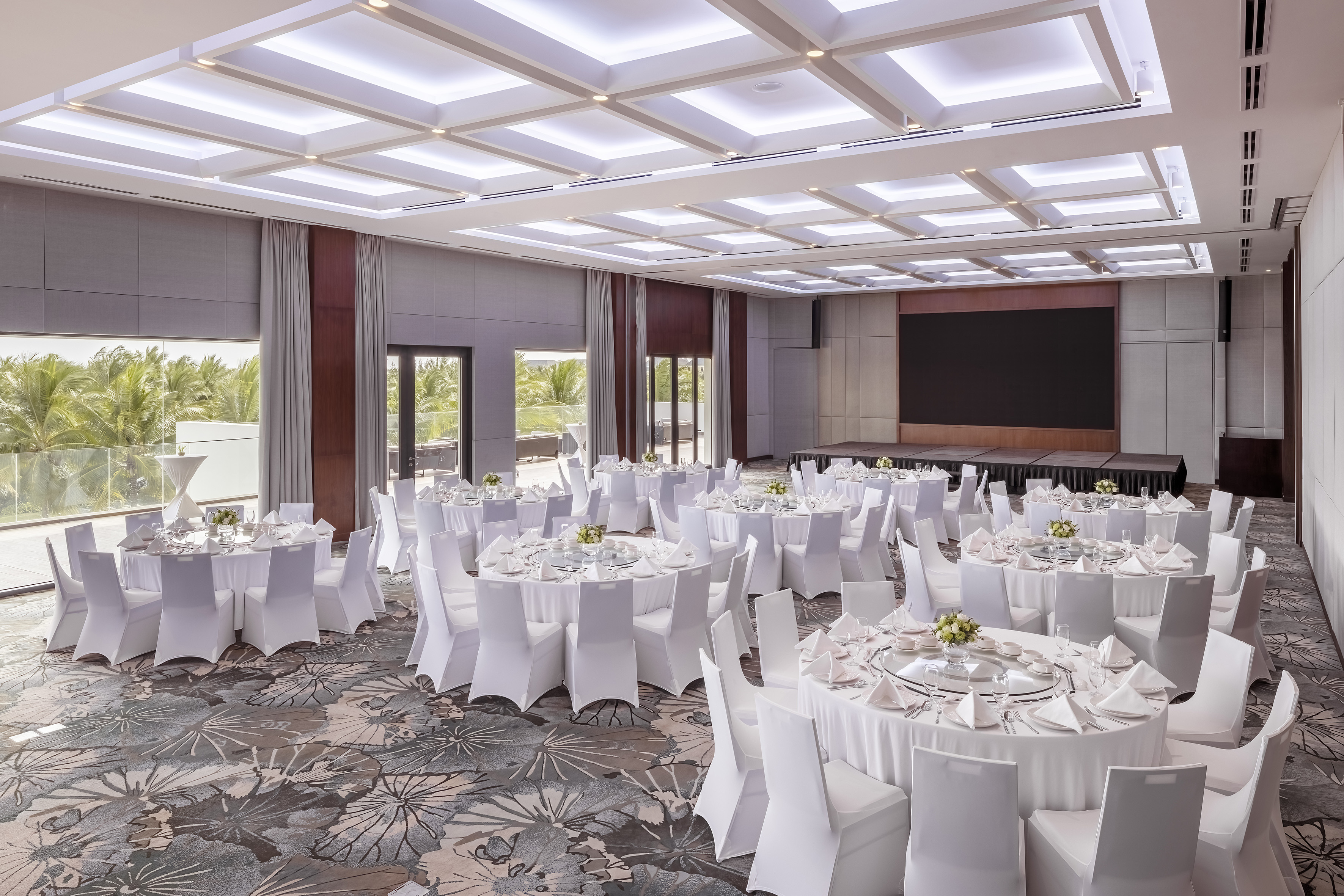 Elegant ballroom with round banquet tables, white chairs, stage, and palm view.