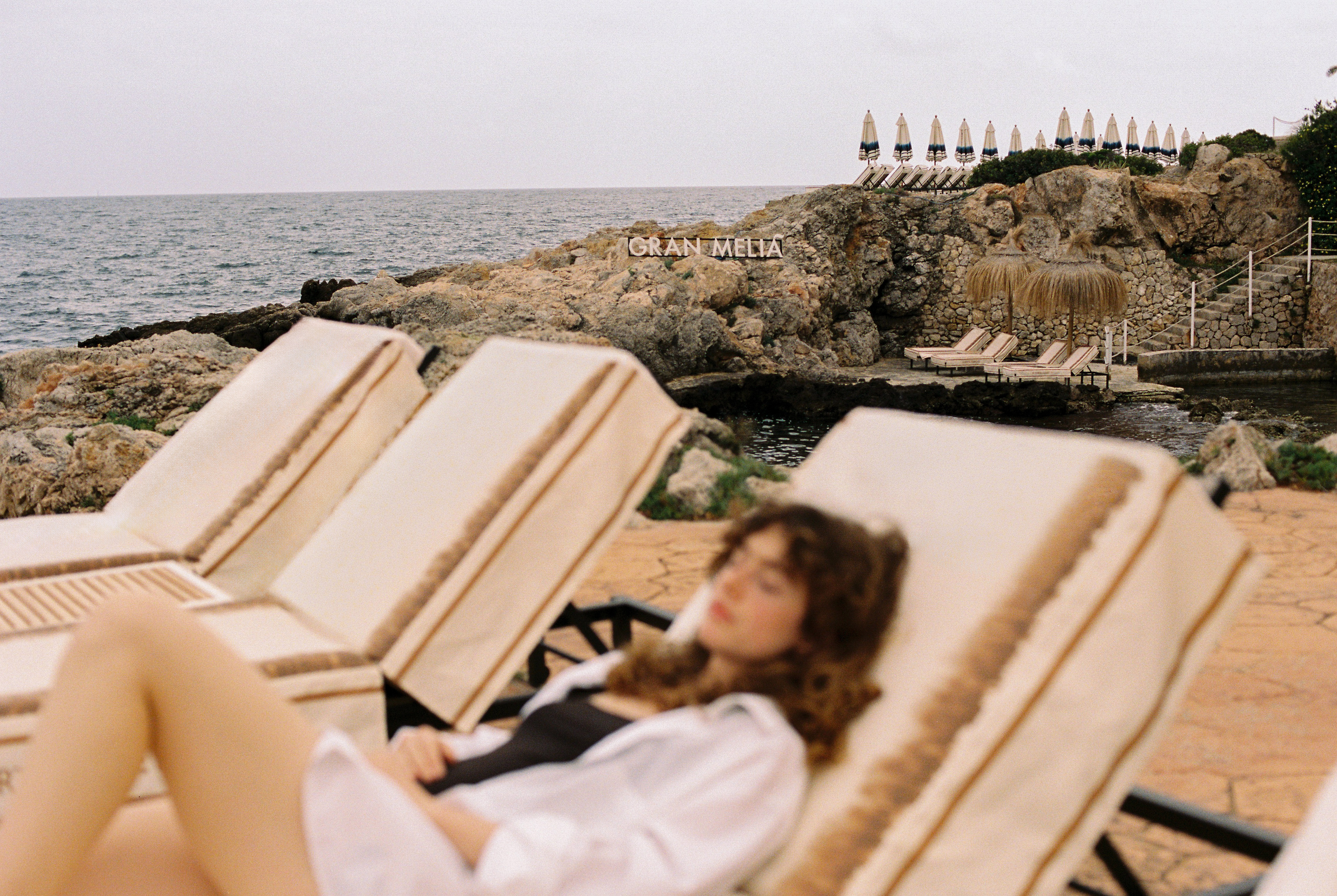 Mujer relajada en tumbona, hotel Gran Meliá en costa rocosa con mar y sombrillas.