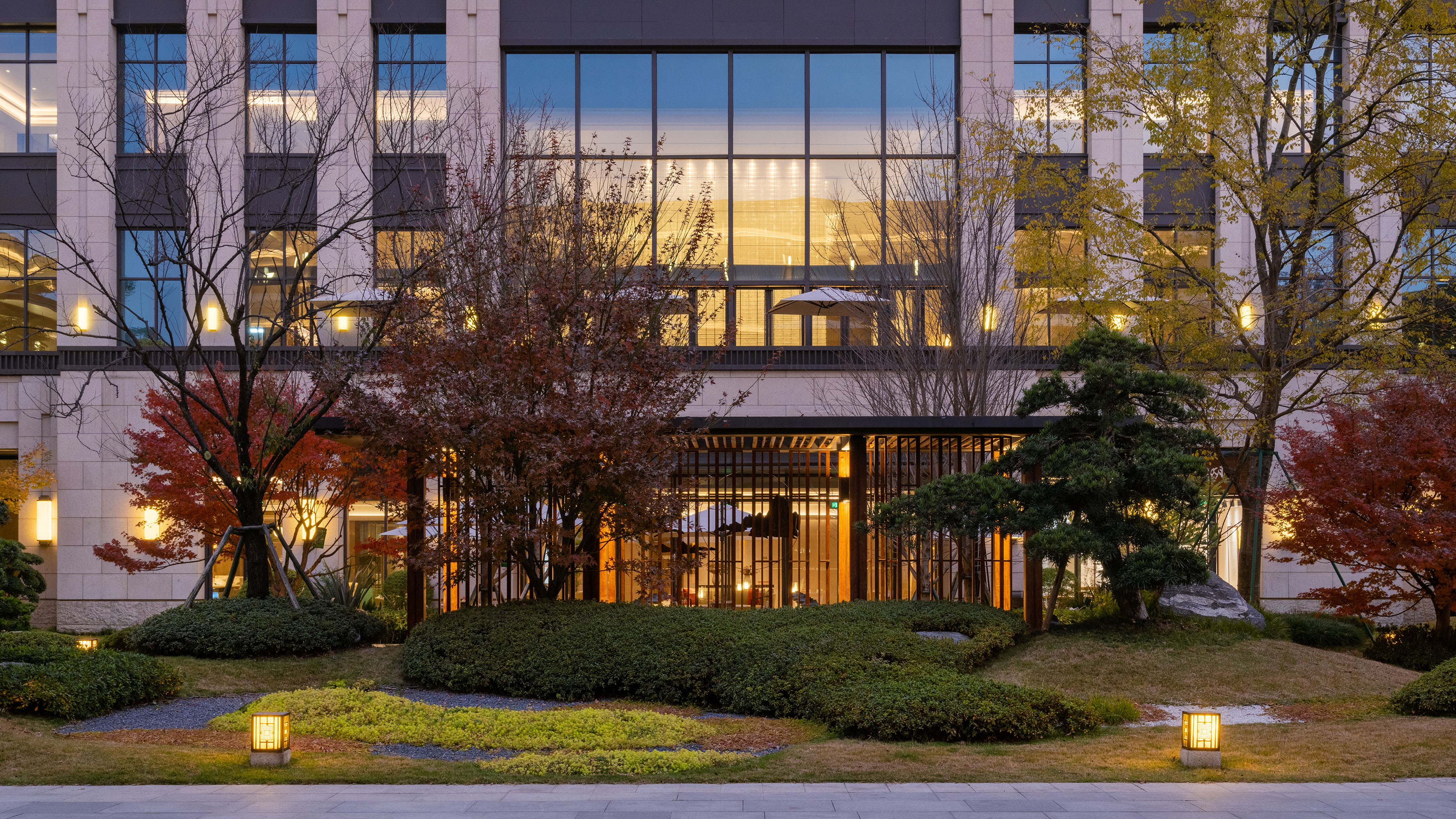 Modern building with autumn trees and a lit landscaped garden.