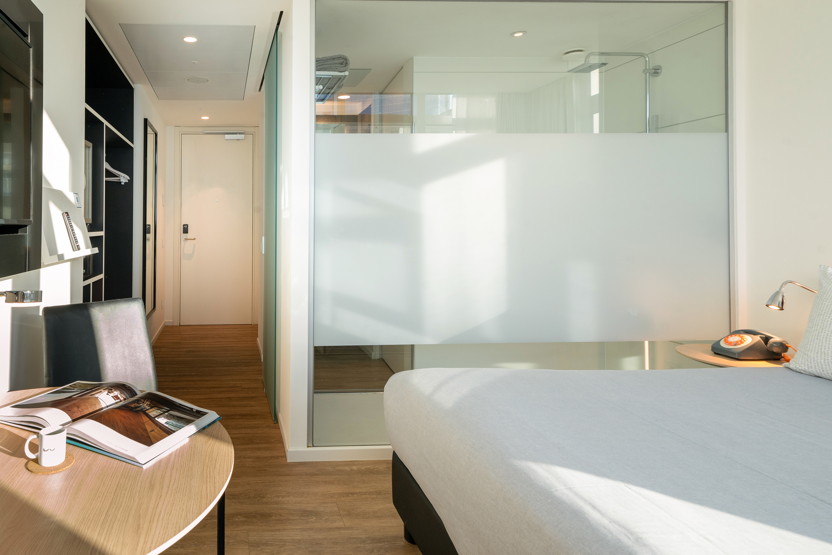 Bright modern hotel room with bed, glass shower, and wooden floor.