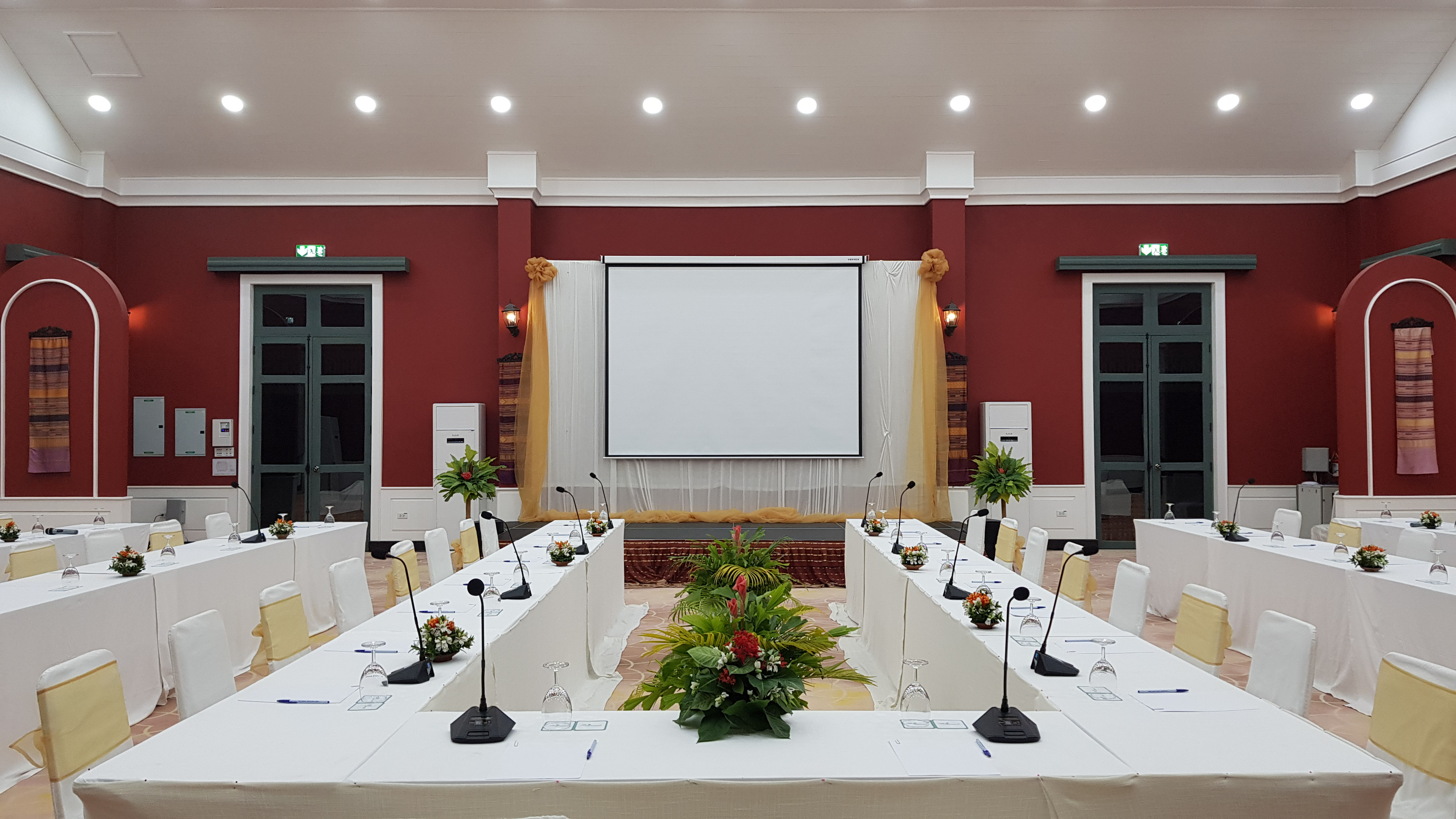 Conference room with U-shaped tables, projector screen, microphones, and red walls.