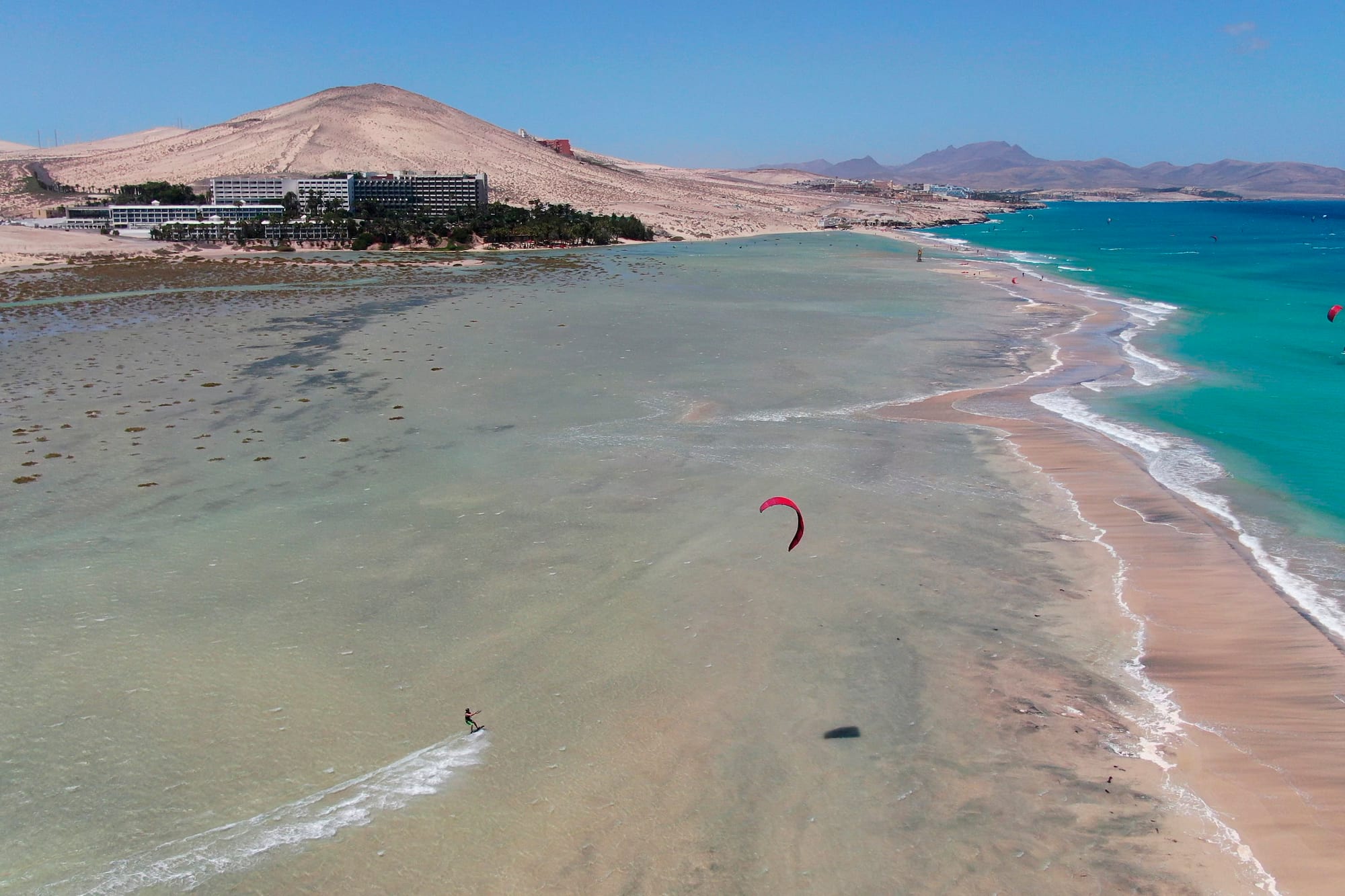 Kitesurfer en playa de arena con aguas turquesas, dunas y hoteles costeros.