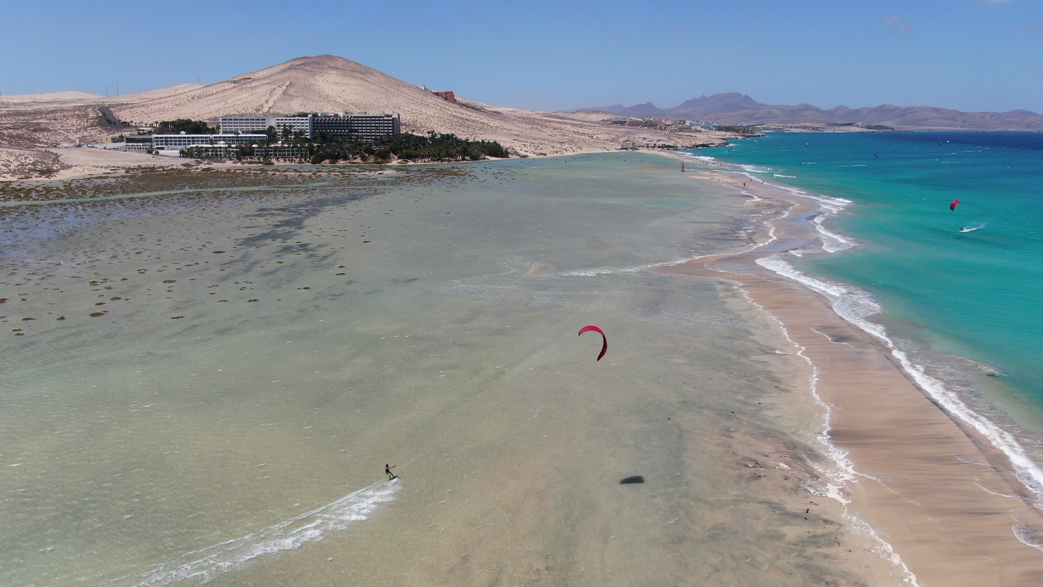 Kitesurfer en playa de arena con aguas turquesas, dunas y hoteles costeros.