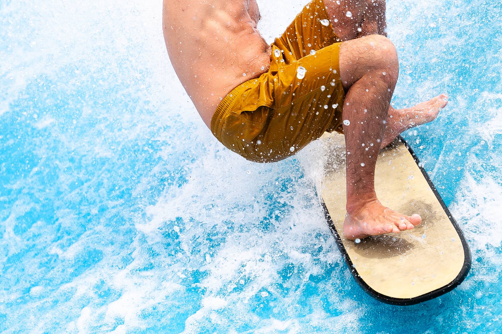 Smiling man flowboarding on a blue artificial wave in yellow shorts.
