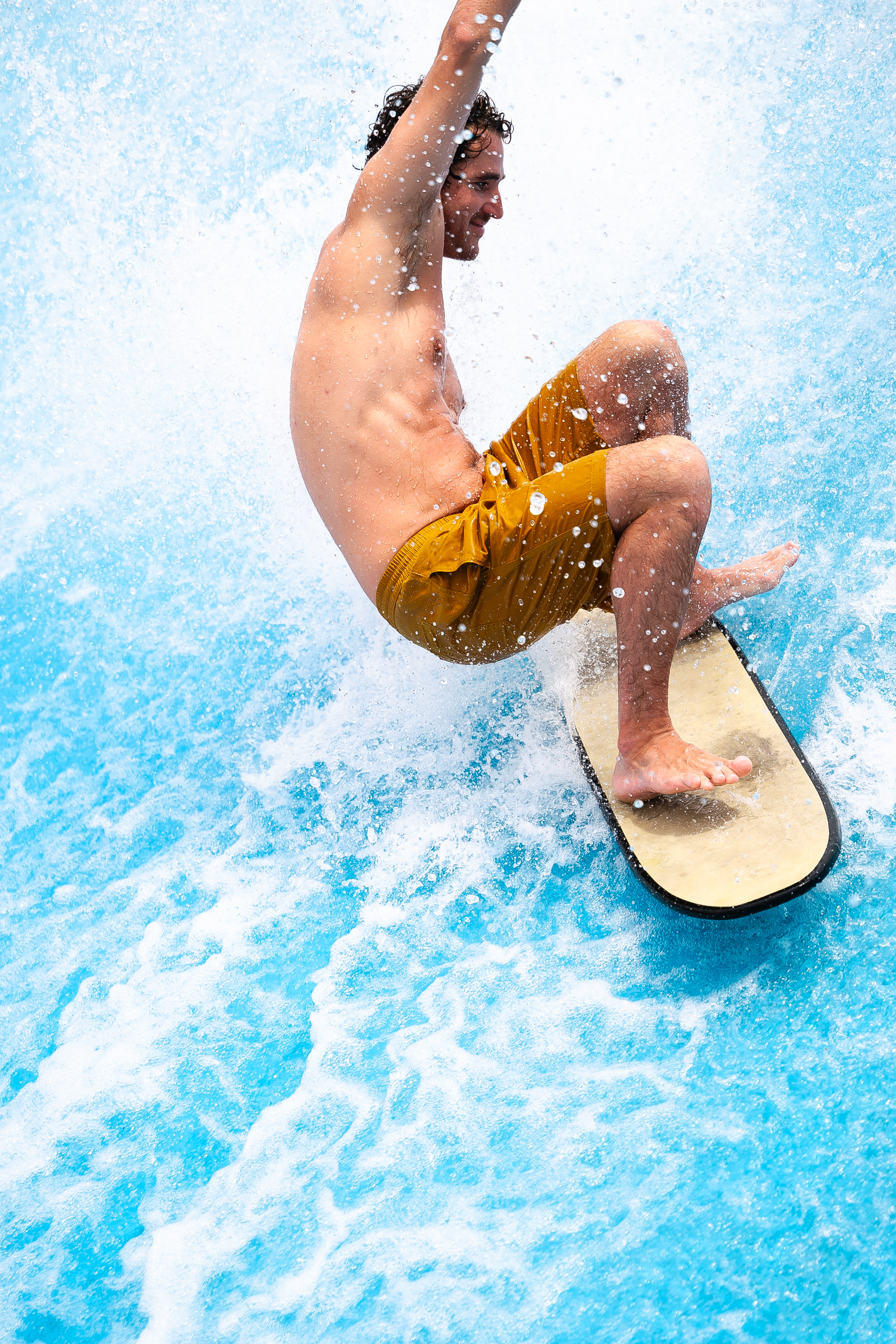 Smiling man flowboarding on a blue artificial wave in yellow shorts.