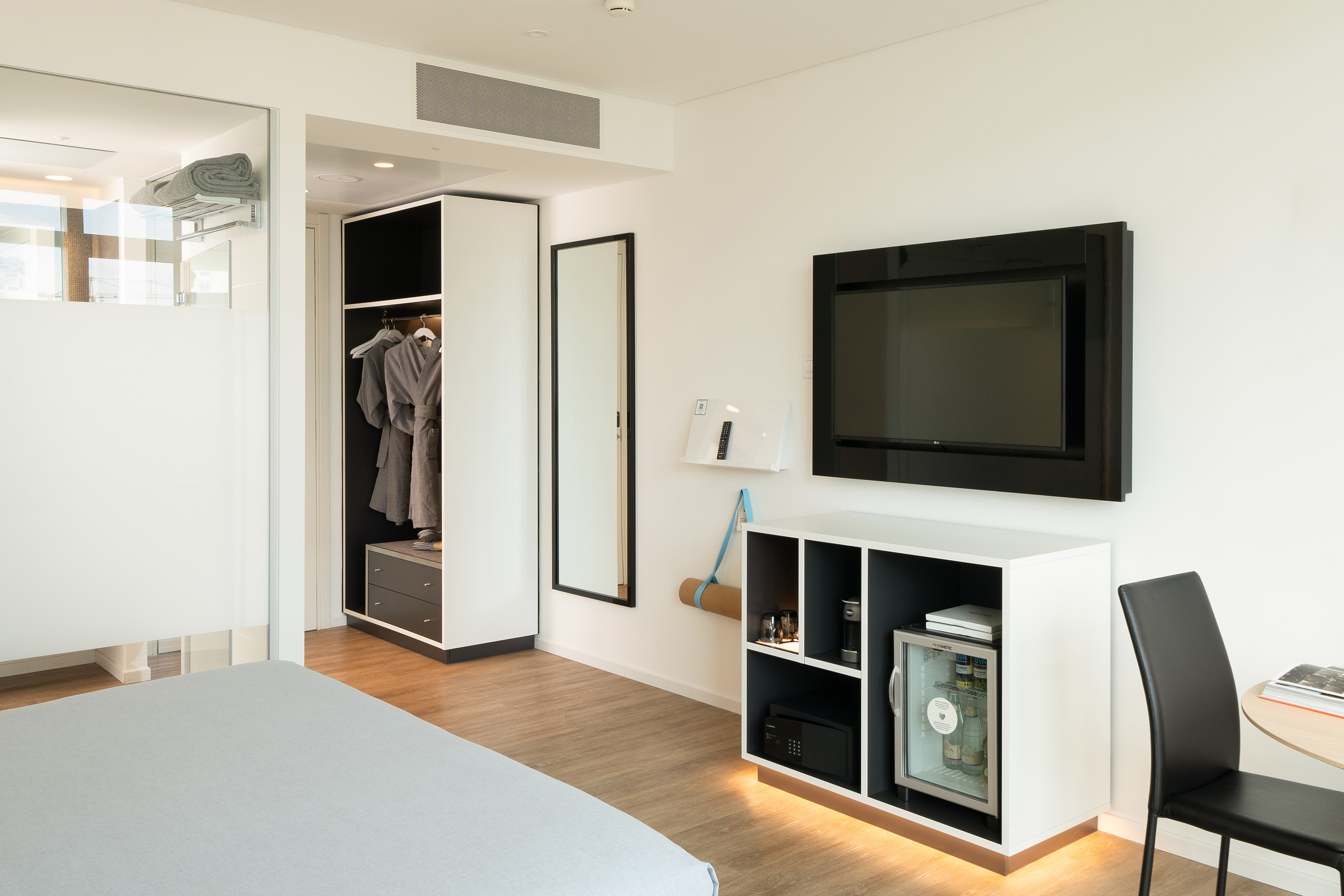 Modern hotel room with wall-mounted TV, minibar, open closet, and yoga mat.
