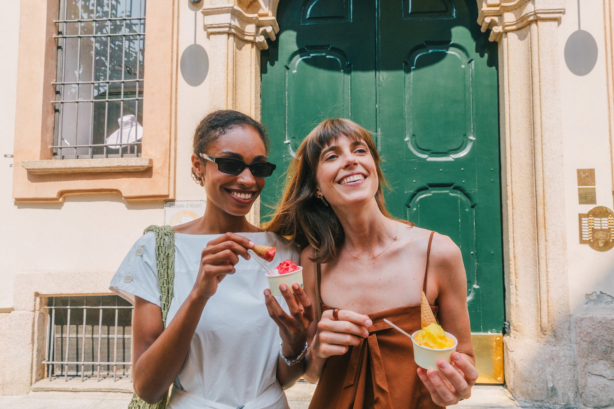 Two smiling female friends eating Italian gelato outside in the sun.
