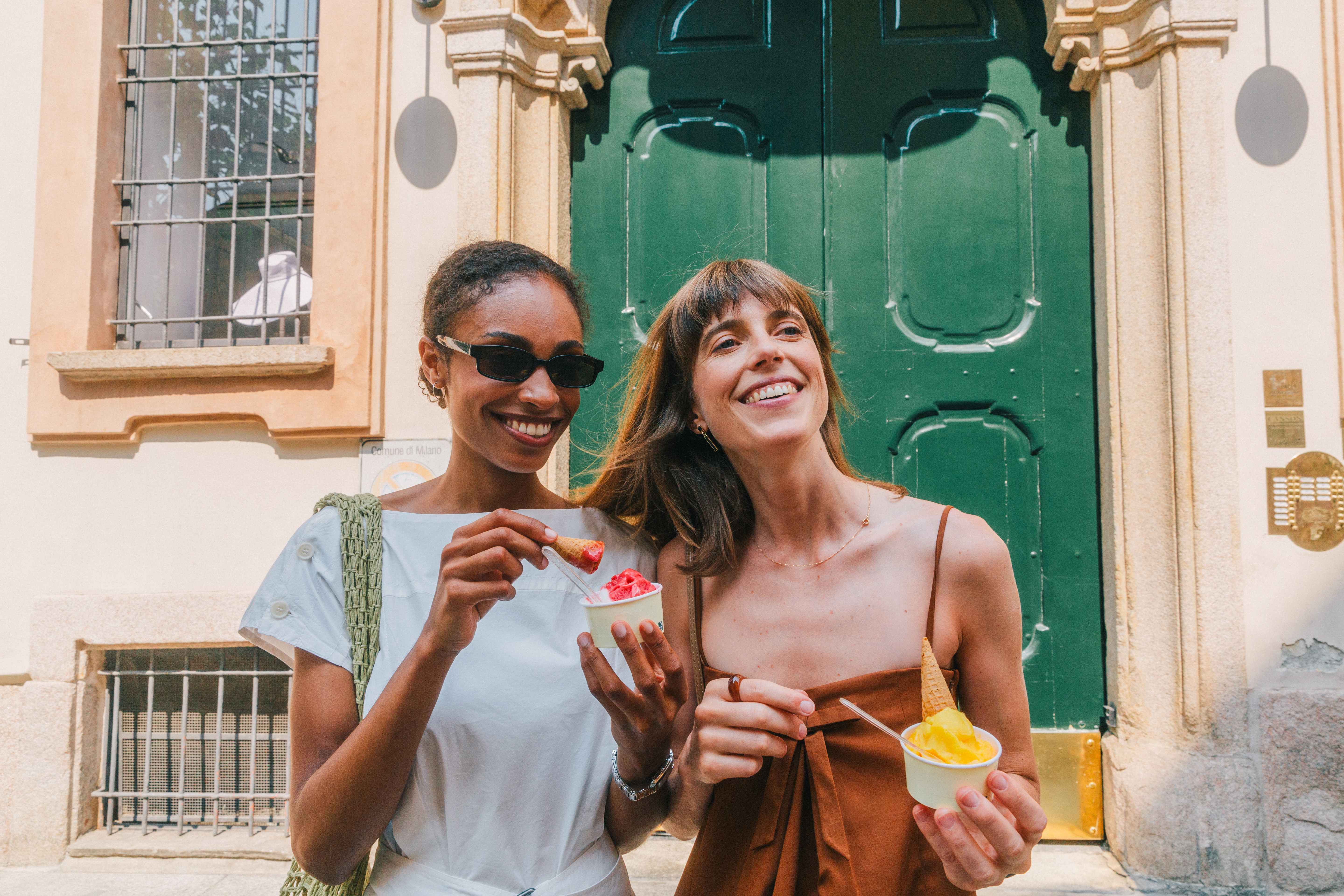 Two smiling female friends eating Italian gelato outside in the sun.