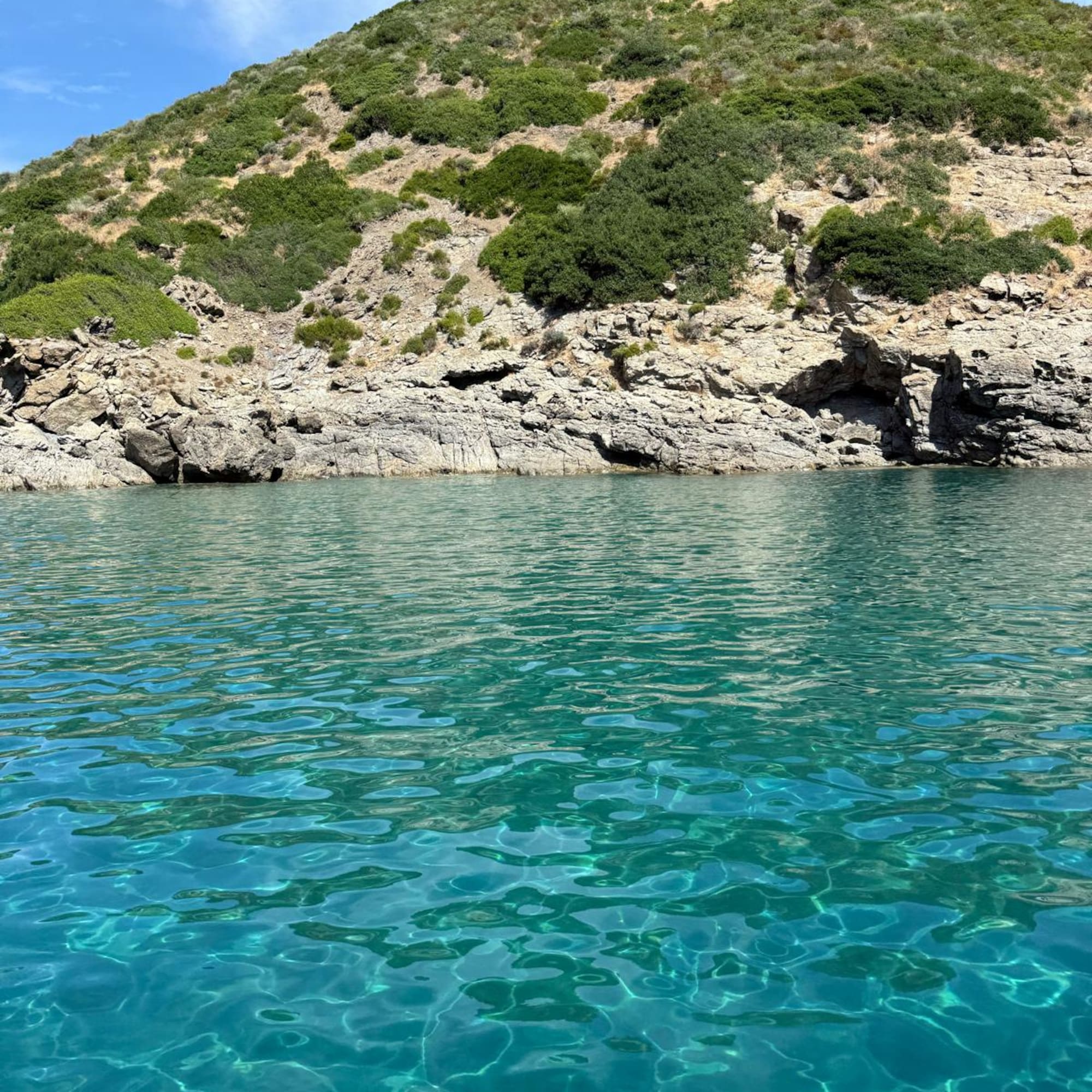 Sparkling turquoise water by a rugged rocky coast and green hillside.